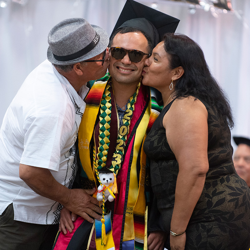 A male graduate gets kissed on the cheek by both his parents during the 2024 Raza graduation.