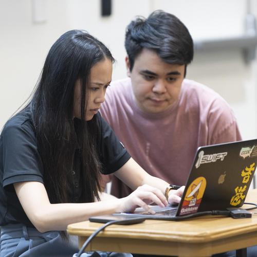 A student using a laptop while another shares her screen inside a classroom.