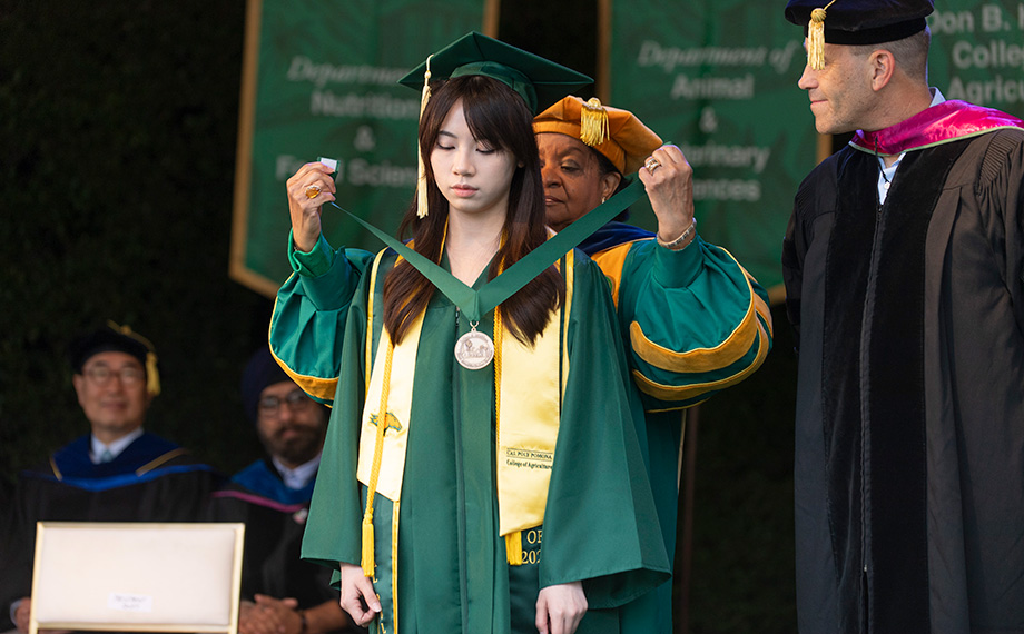 An older woman drapes a medallion around a younger woman's neck while a man watches.