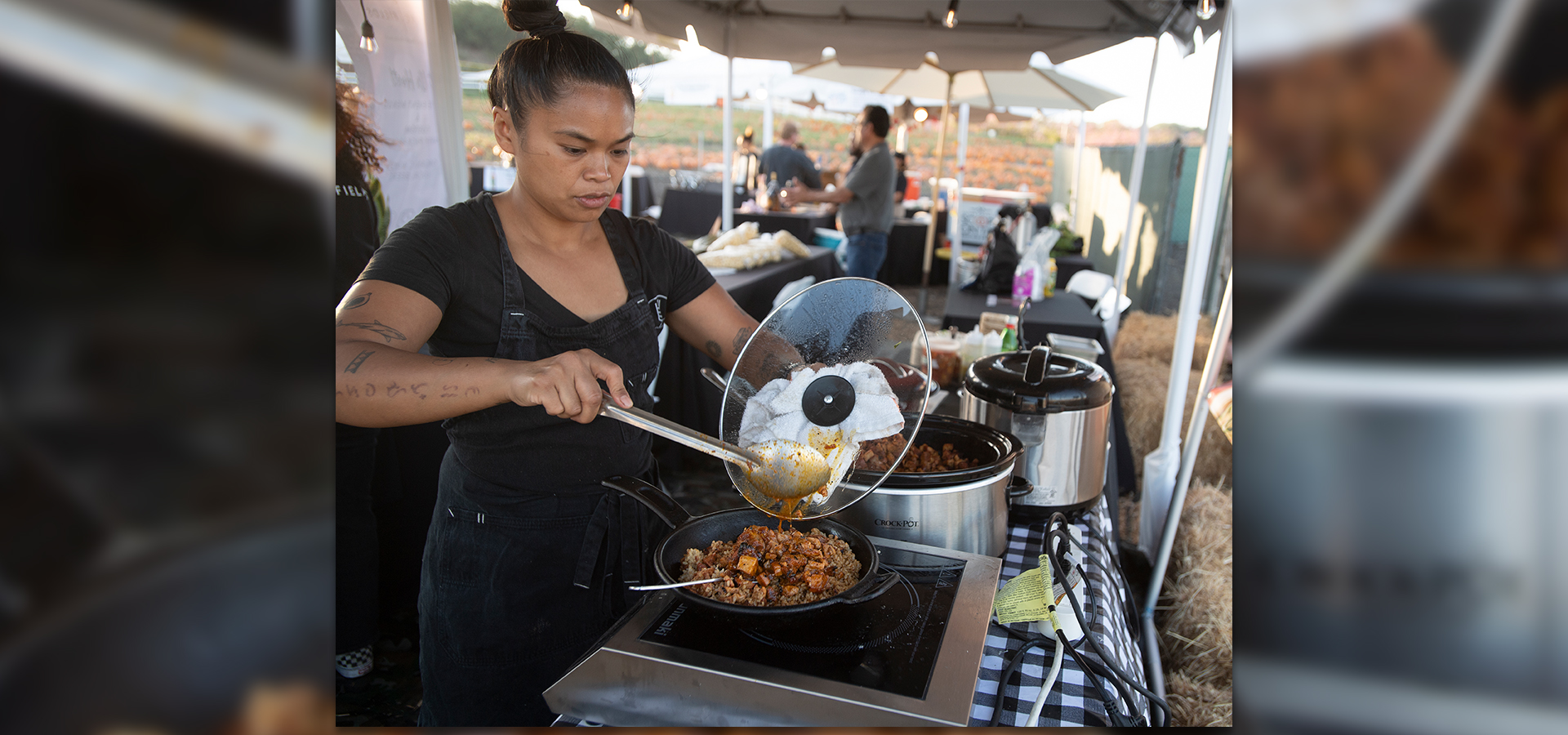 A female cook holds a lid and serving spoon over a frying pan full of food.