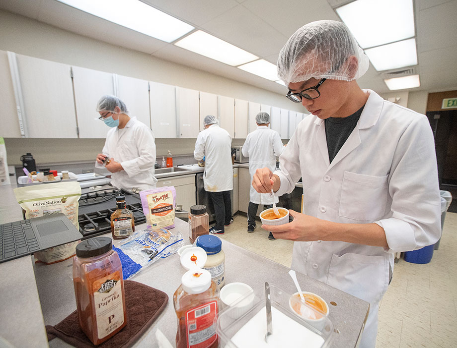 Students in lab coats and head coverings work in a kitchen lab.