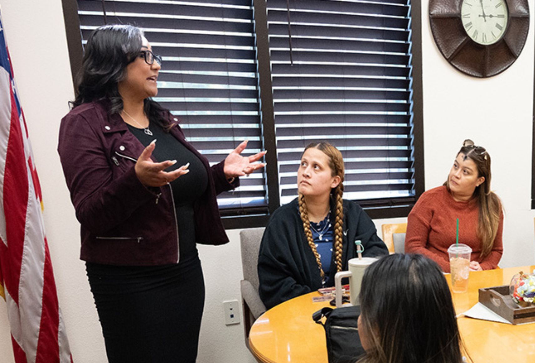 "Four women in a meeting, with one standing and speaking while the others listen."