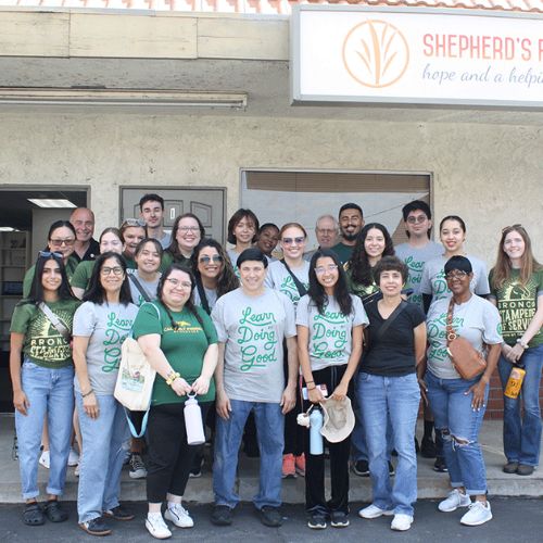 A group of volunteers smiling and posing together in front of a building with a sign that reads "Shepherd's Pantry: hope and a helping hand.