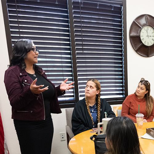 "Four women in a meeting, with one standing and speaking while the others listen."