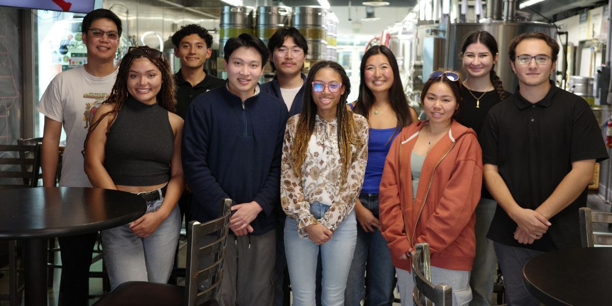 A group of ten young adults standing in a casual indoor setting with brewing equipment in the background, smiling at the camera. The image depicts a diverse and friendly gathering.