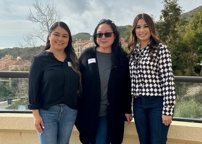 Three women standing on an outdoor balcony, smiling with a scenic backdrop of trees and buildings in the distance.