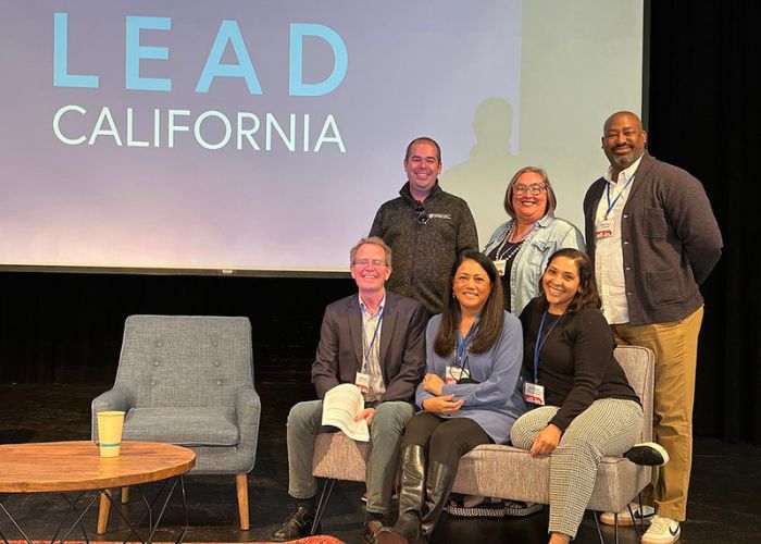 A group of five people posing on stage in front of a large screen that reads "LEAD California," with two seated on chairs and three standing behind them.