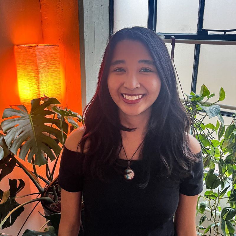 Yna Gatpandan a young woman with long black hair in an off-shoulder black top, smiling in a cozy room with plants and warm lighting.