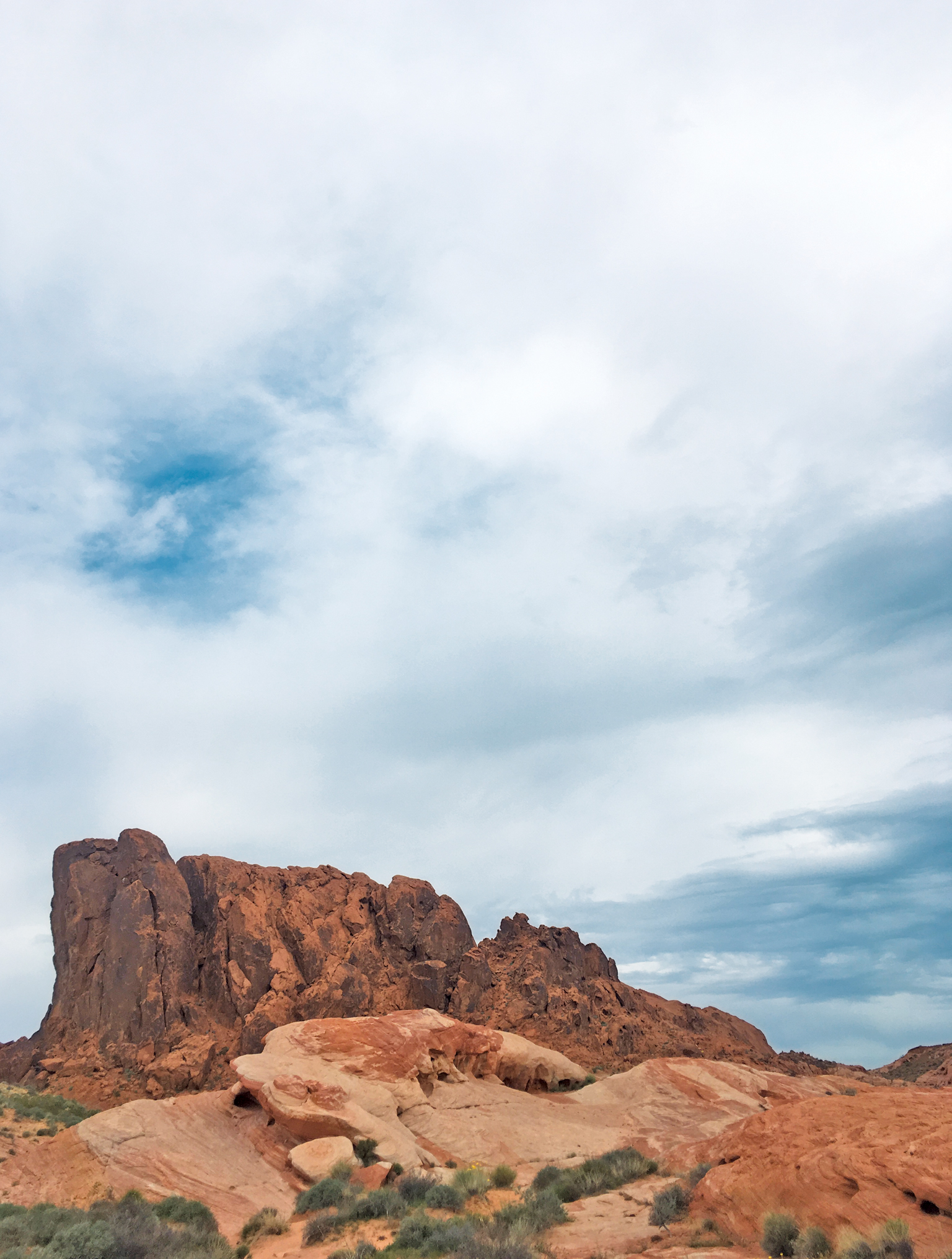 A rugged desert scene under a cloudy sky. Red rock formations rise sharply in the middle of the landscape, their rough surfaces contrasting with the softer clouds above.