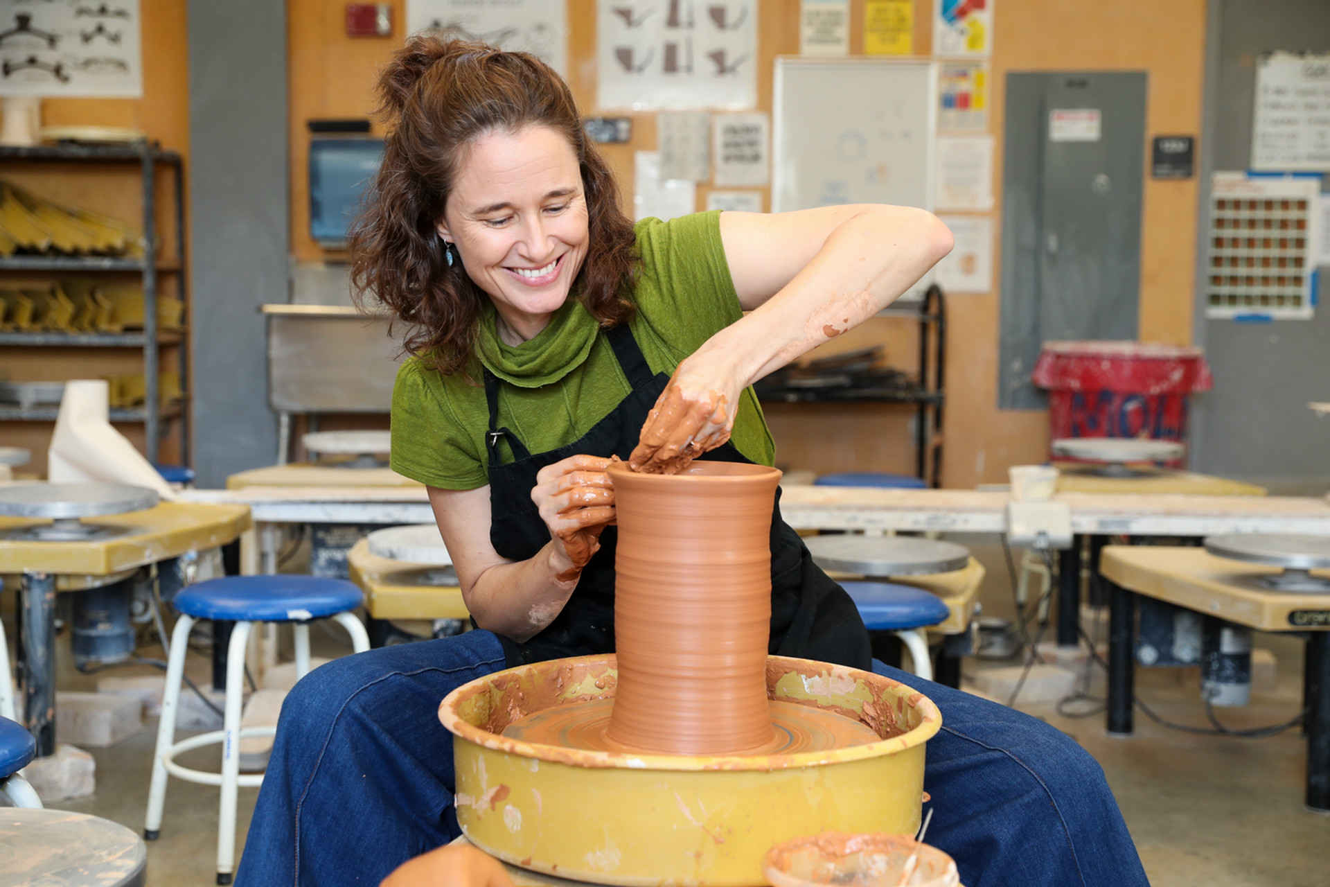 Alison Ragguette, she is smiling happily whilewearing a green shirt with a black apron as she sculpts clay using a pottery wheel.