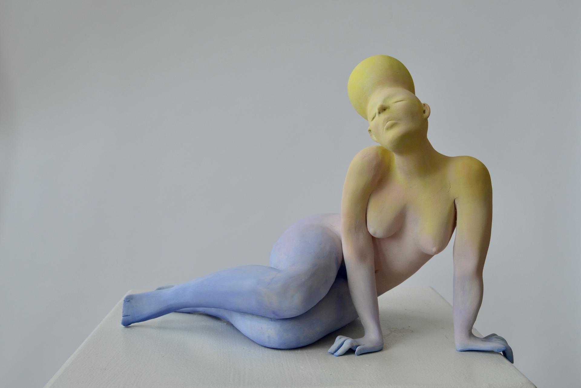 a sculpture of a nude, abstracted female figure in a seated, reclining pose. The figure’s body appears to be gracefully contorted, with one leg extended and the other bent beneath the body. One arm is bent at the elbow, supporting the figure as it leans back, while the other arm rests casually on the surface. The head is tilted upward, with eyes closed and lips gently pressed together, giving the figure a calm, serene expression.