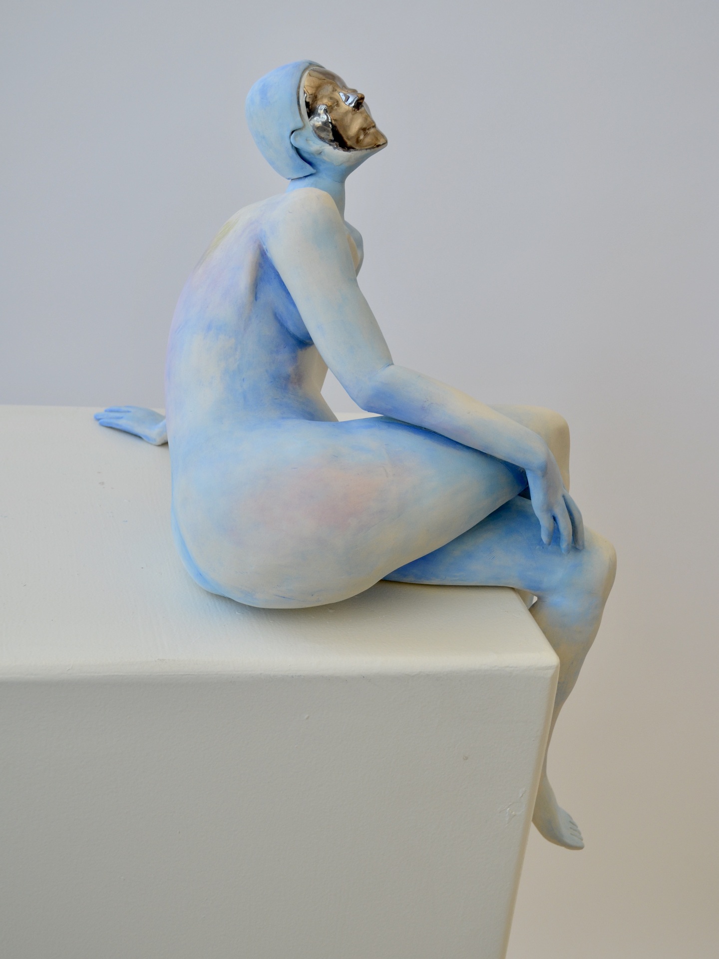 a sculpture of a seated, nude figure positioned at the edge of a flat surface. The figure is leaning back slightly, with one arm resting behind for support, while the other arm rests on the bent knee. The sculpture is smooth, with a light, soft color palette of pale blues and subtle pinks, giving the body a cool, ethereal appearance.