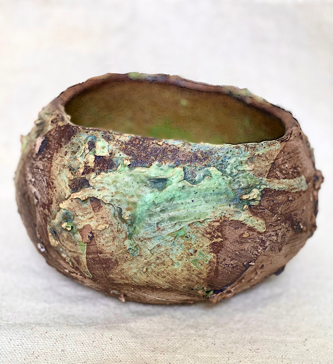 A handcrafted, rustic ceramic bowl with a rough, organic texture. The exterior surface of the bowl is predominantly brown with patches of green glaze that appear to have been applied unevenly, creating an earthy, weathered look. The green glaze is concentrated in certain areas, blending naturally with the brown tones of the clay.