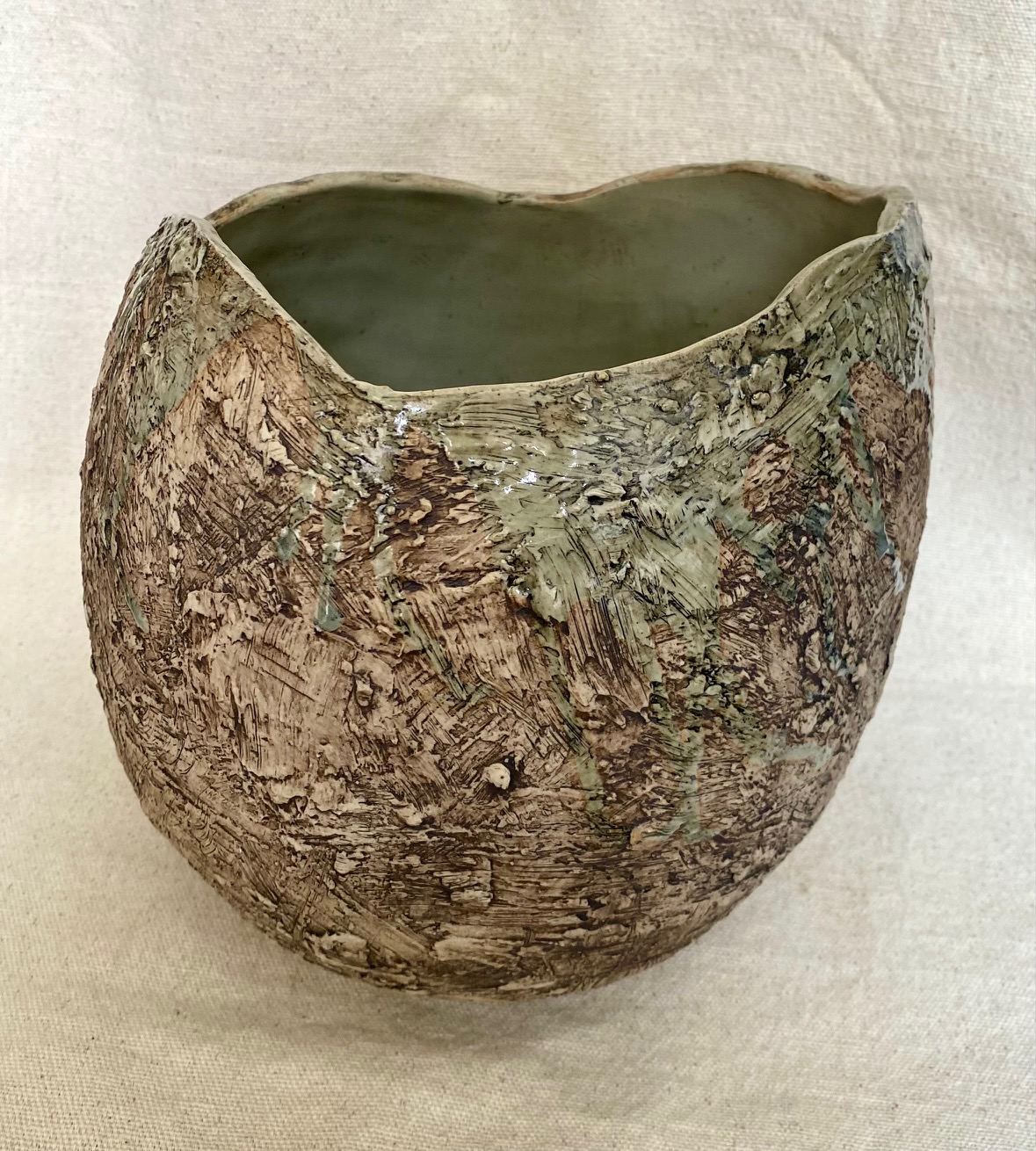 A handcrafted ceramic vessel with an organic, earthy texture. The exterior of the vessel is rough, with a mix of brown and green tones, resembling a natural, weathered surface. The surface appears to have been textured with wide, sweeping marks and patches, adding a sense of depth and irregularity to the form. There are subtle hints of green glaze applied in uneven areas, which blend into the natural brown clay, giving the piece a moss-like appearance.