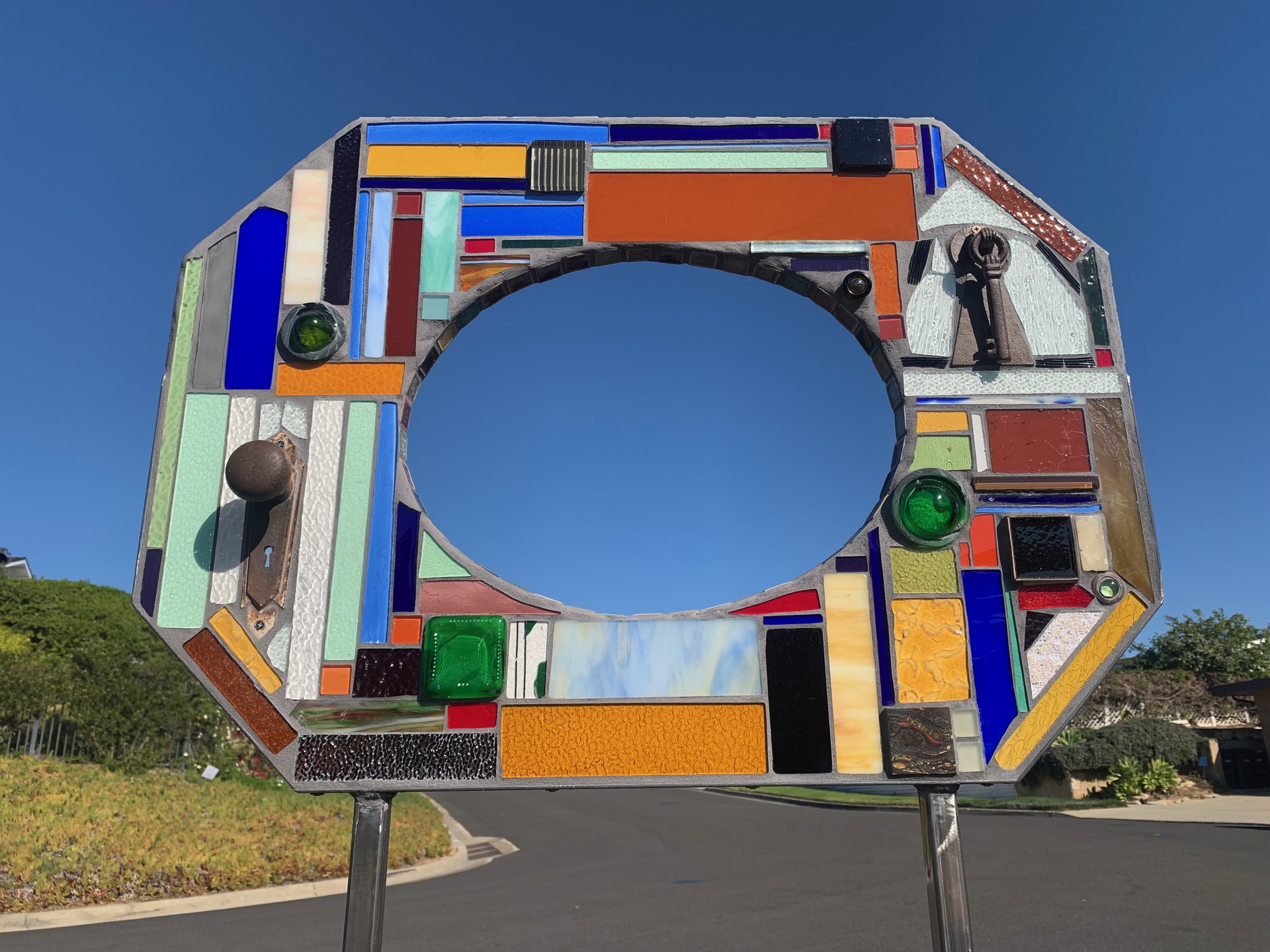 An octagonal stained glass artwork, consisting of a variety of vibrant, rectangular glass panes and other found objects. The center is open, creating a window-like effect within the frame. The glass pieces are arranged in a mosaic, featuring a range of colors including blues, oranges, greens, and yellows, interspersed with textured or translucent glass sections.