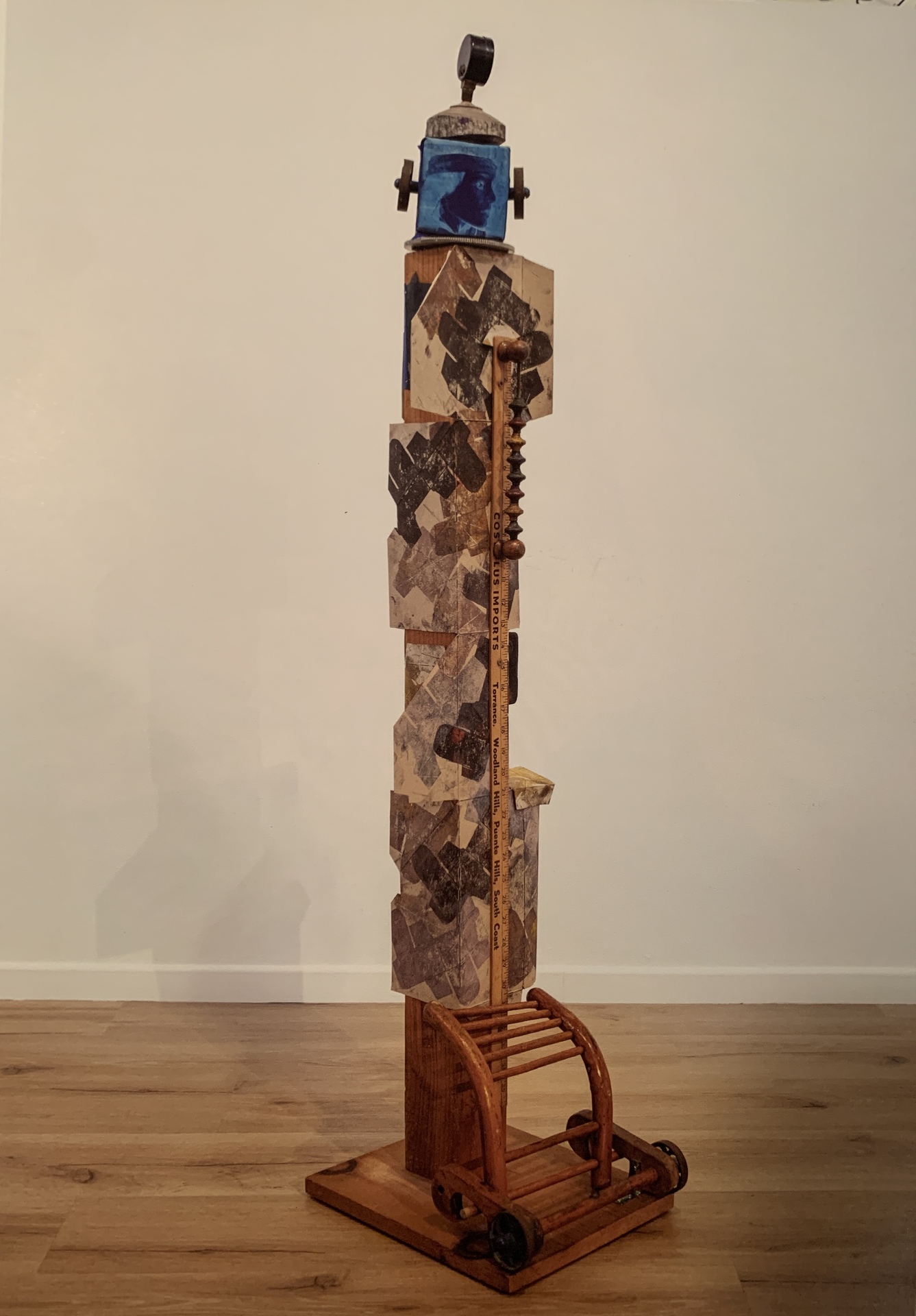 A tall, abstract wooden sculpture composed of various materials and geometric shapes. The base is a wooden platform, supporting a vertical structure that includes a mix of natural and industrial elements. At the bottom of the sculpture, there is a small wooden component with wheels, resembling a miniature cart or trolley.