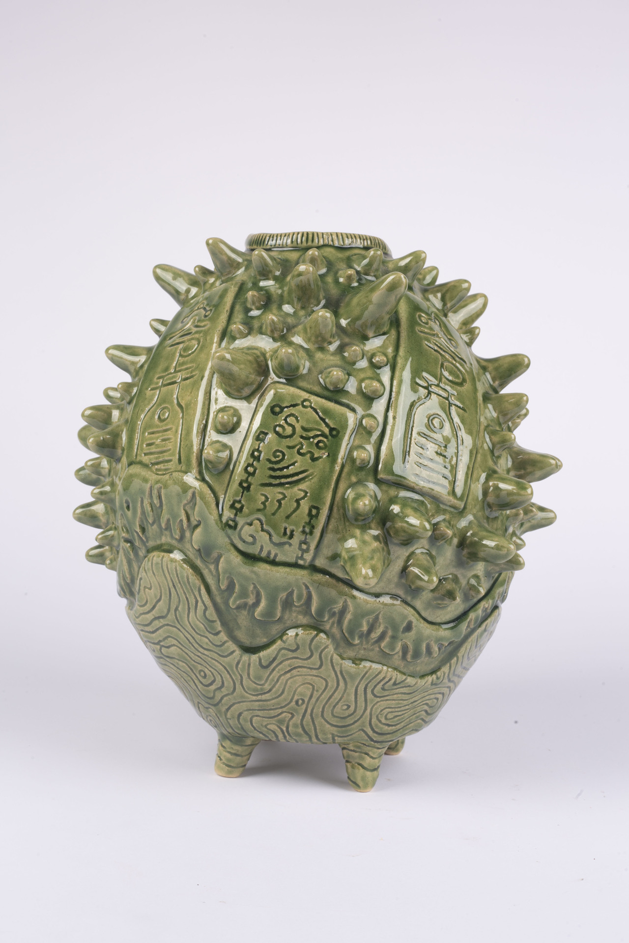 A unique, green-glazed ceramic vessel with an irregular, spherical shape. The surface of the vessel is covered with a combination of sharp, spiky protrusions and smooth, carved designs. The spikes vary in size, creating a textured, almost alien appearance. In between the spikes, there are rectangular panels with intricate, abstract carvings that resemble symbols or hieroglyphs, adding a sense of mystery and craftsmanship to the piece.