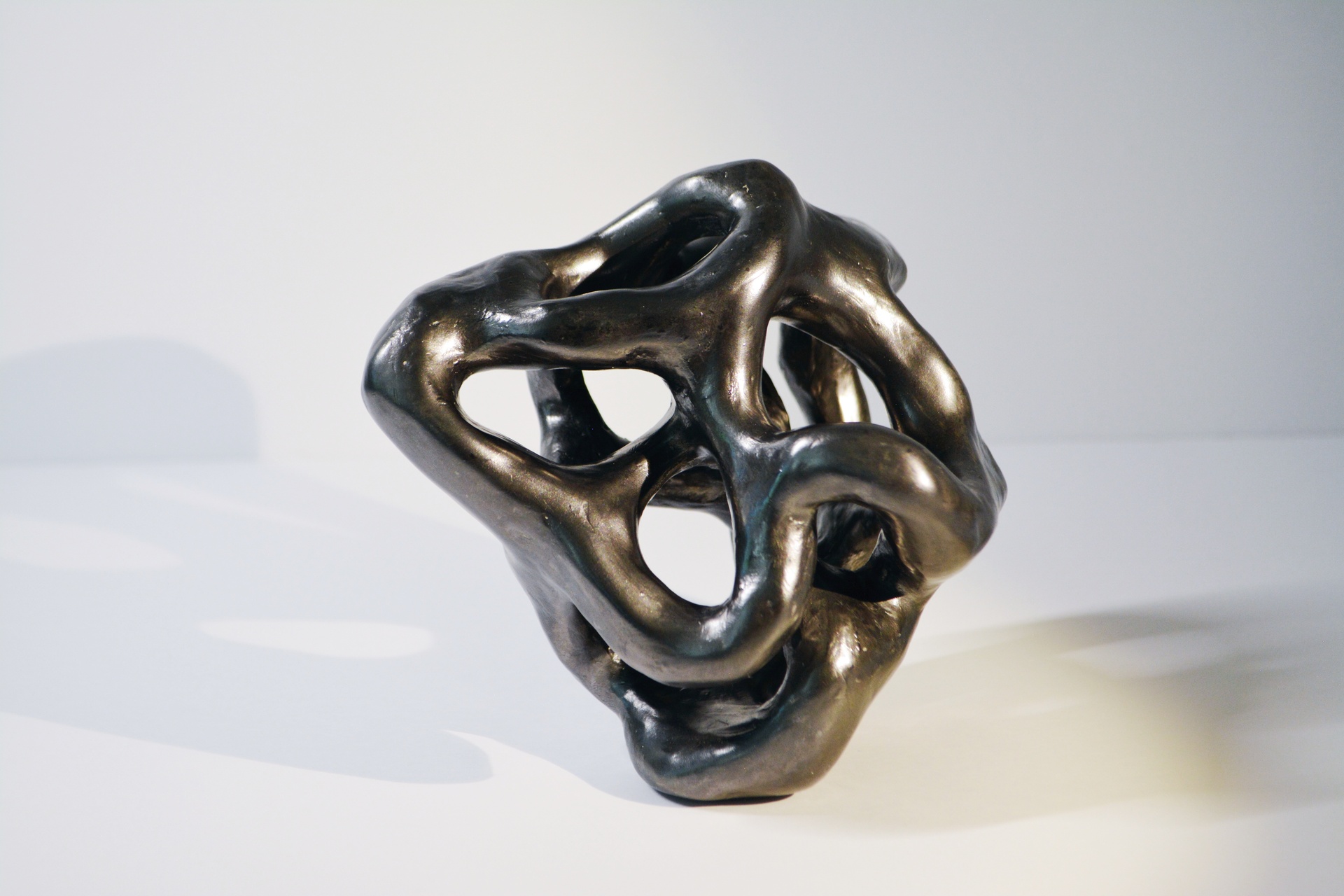 A smooth, abstract bronze sculpture with a flowing, organic form. The piece consists of interwoven, curving shapes that loop and twist into an open, freeform structure. The smooth, polished surface of the bronze reflects light, highlighting the contours and negative spaces between the loops.