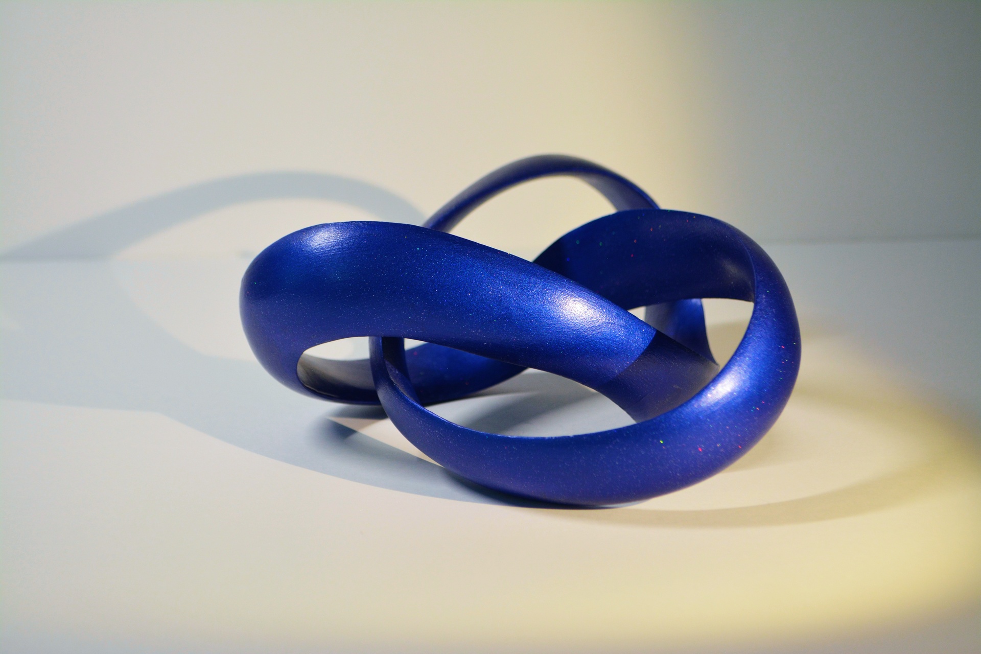 A smooth, abstract sculpture made of interlocking, curved forms that twist and fold into one another. The piece is a deep, rich blue color, with a subtle, speckled finish that adds a hint of texture and shimmer to the surface. The interconnected loops create a sense of continuous motion, and the clean lines and glossy surface give the sculpture a modern, elegant aesthetic.