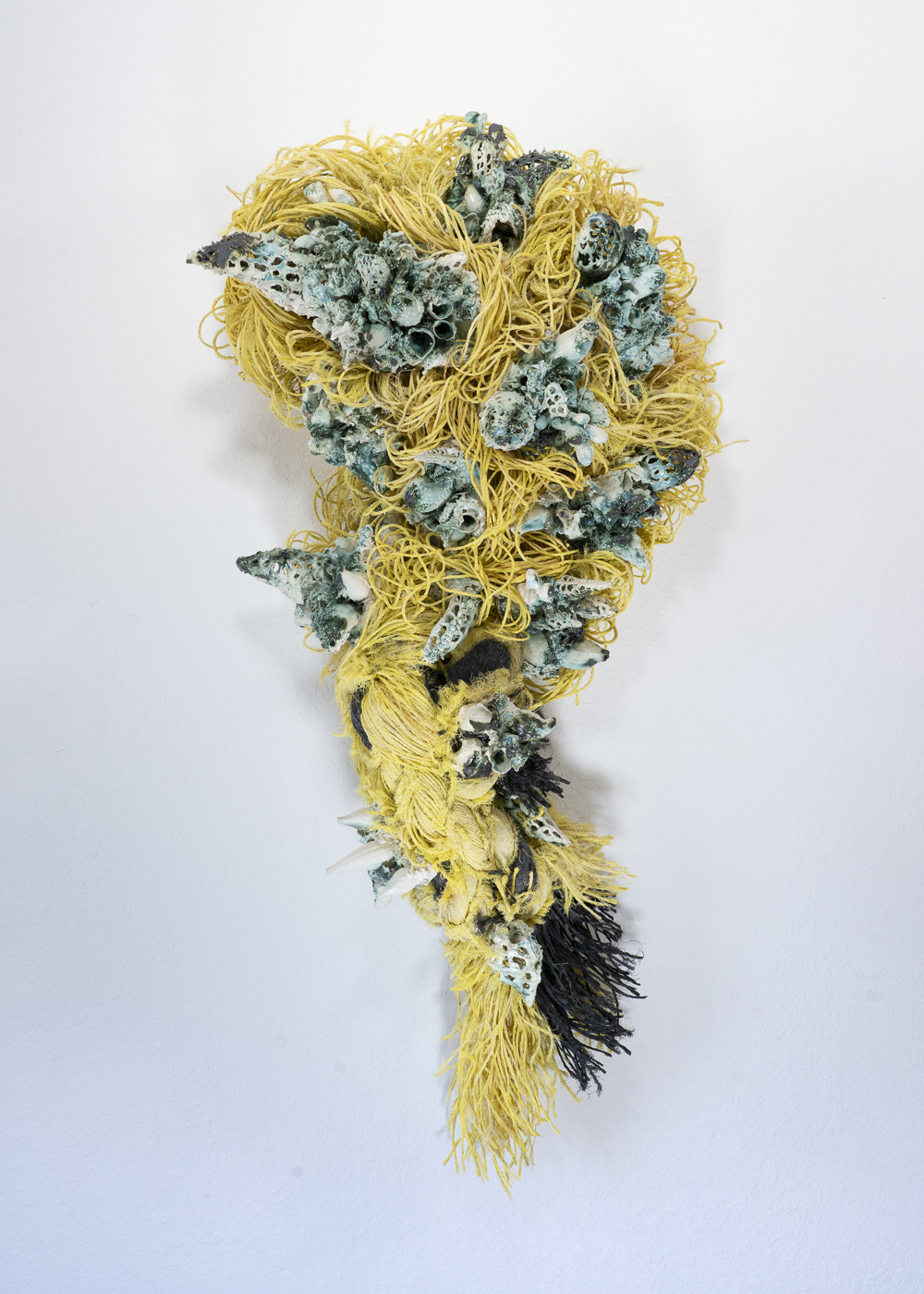 A sculptural wall piece made from a combination of organic and textured materials. The artwork features a mass of tangled yellow fibers, resembling yarn or rope, cascading down in a twisting, flowing form. Embedded within the fibers are intricate ceramic elements, some of which resemble coral or spiky, oceanic structures.