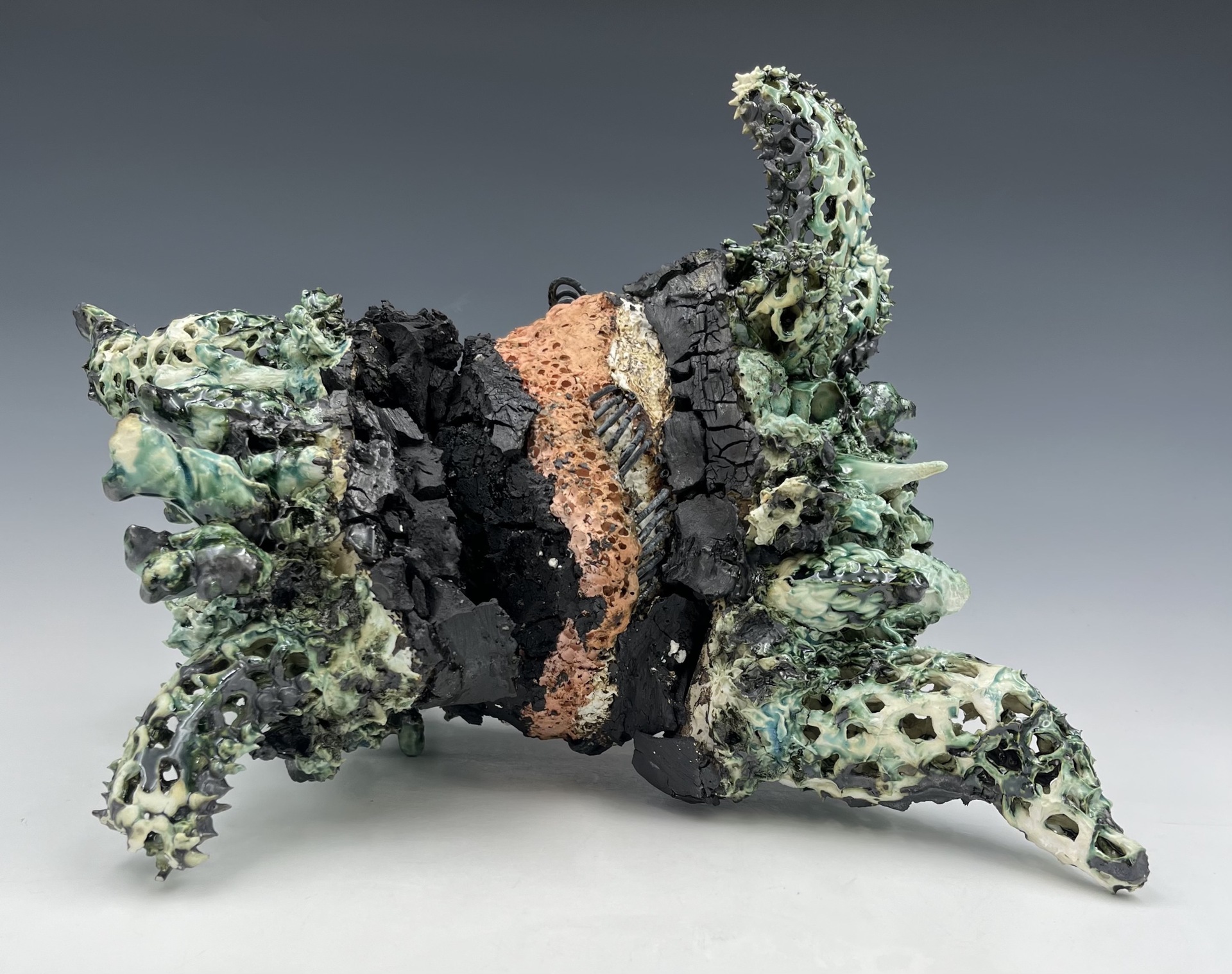 A textured, abstract ceramic sculpture that appears to mimic organic, geological, or coral-like forms. The piece is irregular in shape, with rough, jagged surfaces and a variety of spiked, horn-like protrusions that extend outward in different directions. The colors are primarily muted greens, blacks, and earthy browns, with a blend of glossy and matte finishes that emphasize the contrasts in texture.