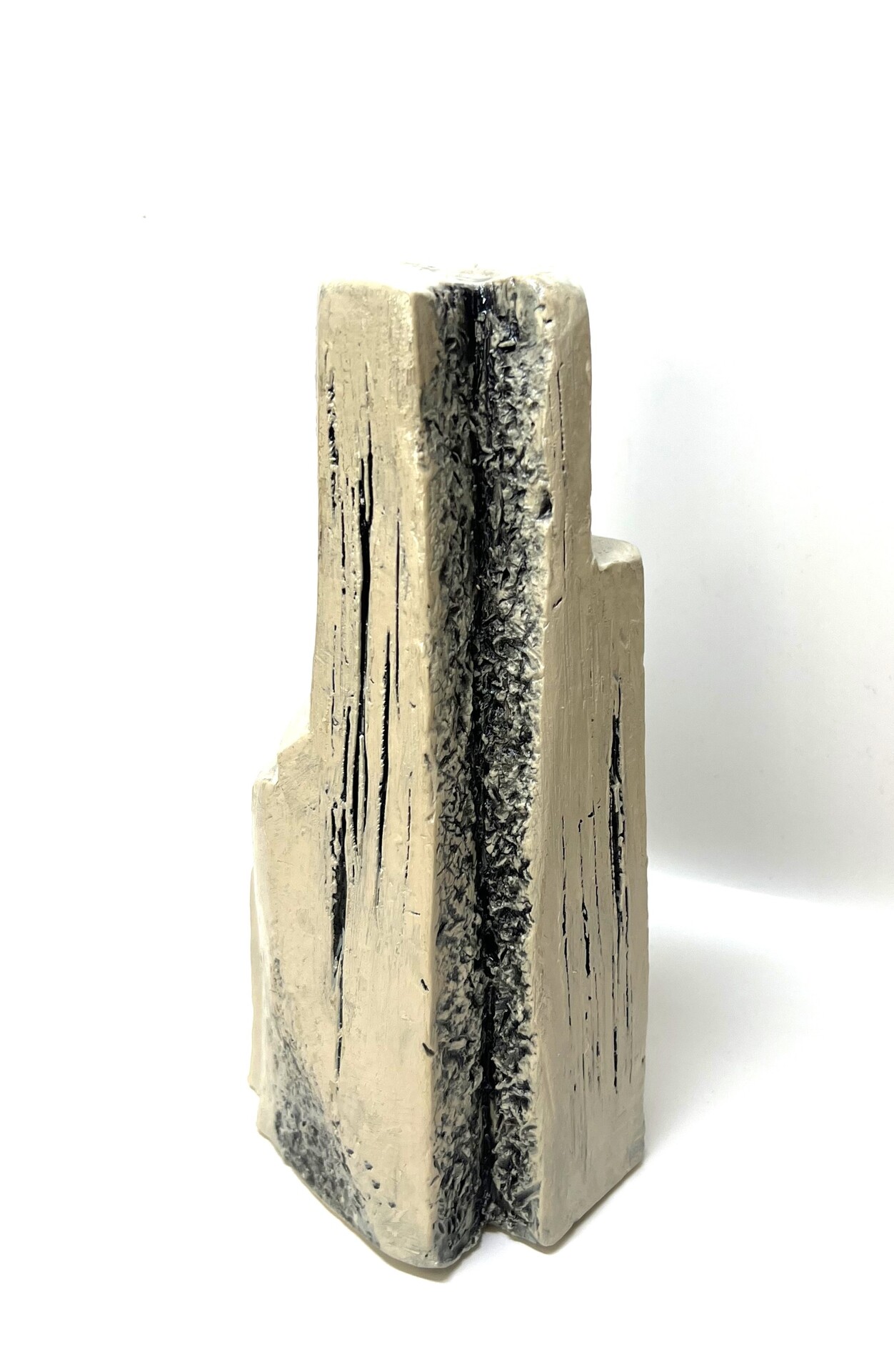 This is a ceramic sculpture with a tall, vertical form. The surface features textured, uneven grooves with a rough, dark crack running down the center. The piece is mostly beige, with areas of black highlighting the rugged texture. The overall shape is blocky and abstract.