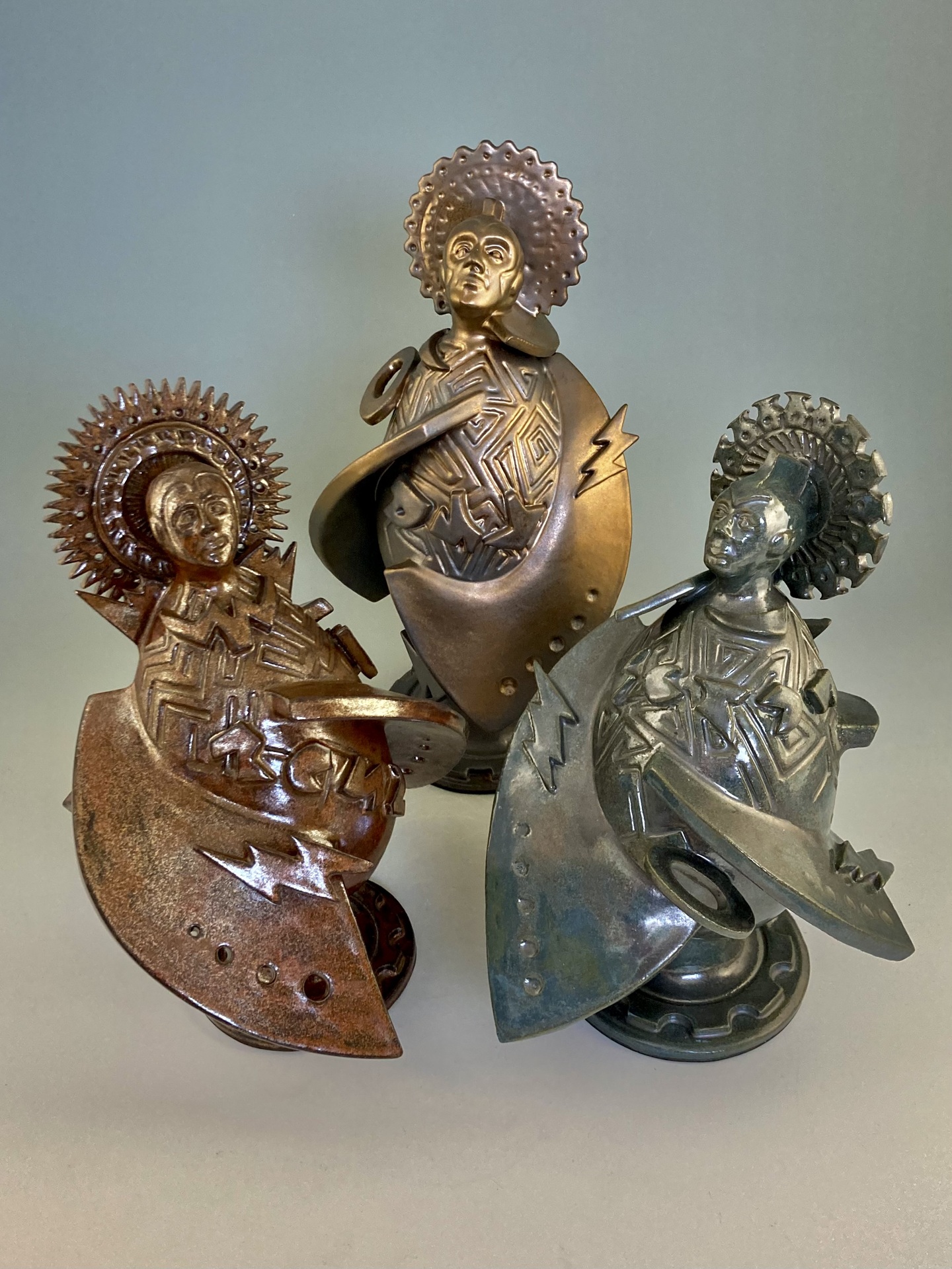 Three small, metallic sculptures featuring figures with intricate geometric patterns on their cloaks and halo-like headpieces, each in a different metallic finish—copper, bronze, and silver.