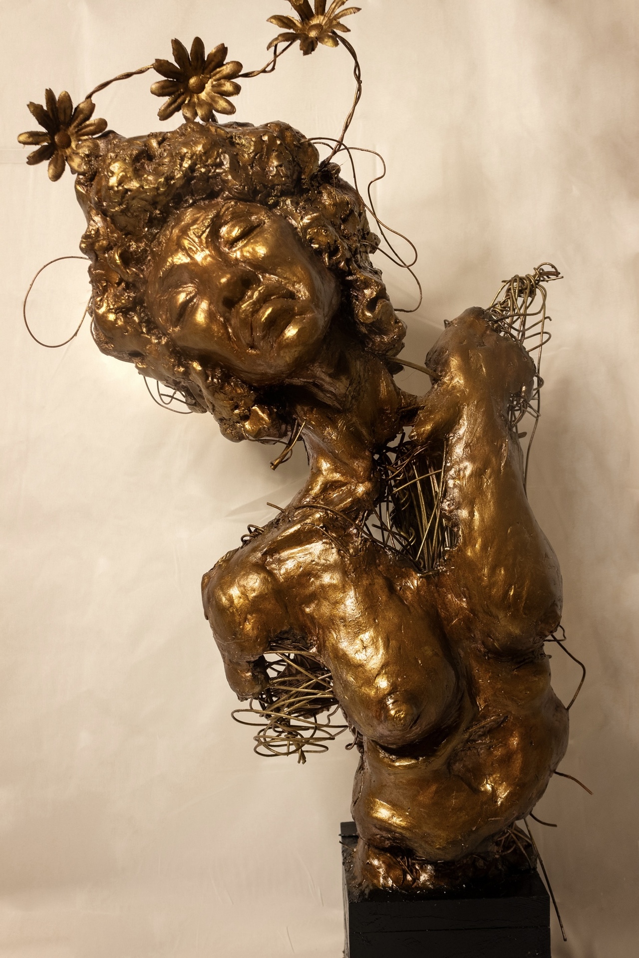  A bronze-colored sculpture of a figure with a peaceful expression, adorned with wire-like details and flowers extending from their head, with parts of the body appearing incomplete or unraveling.