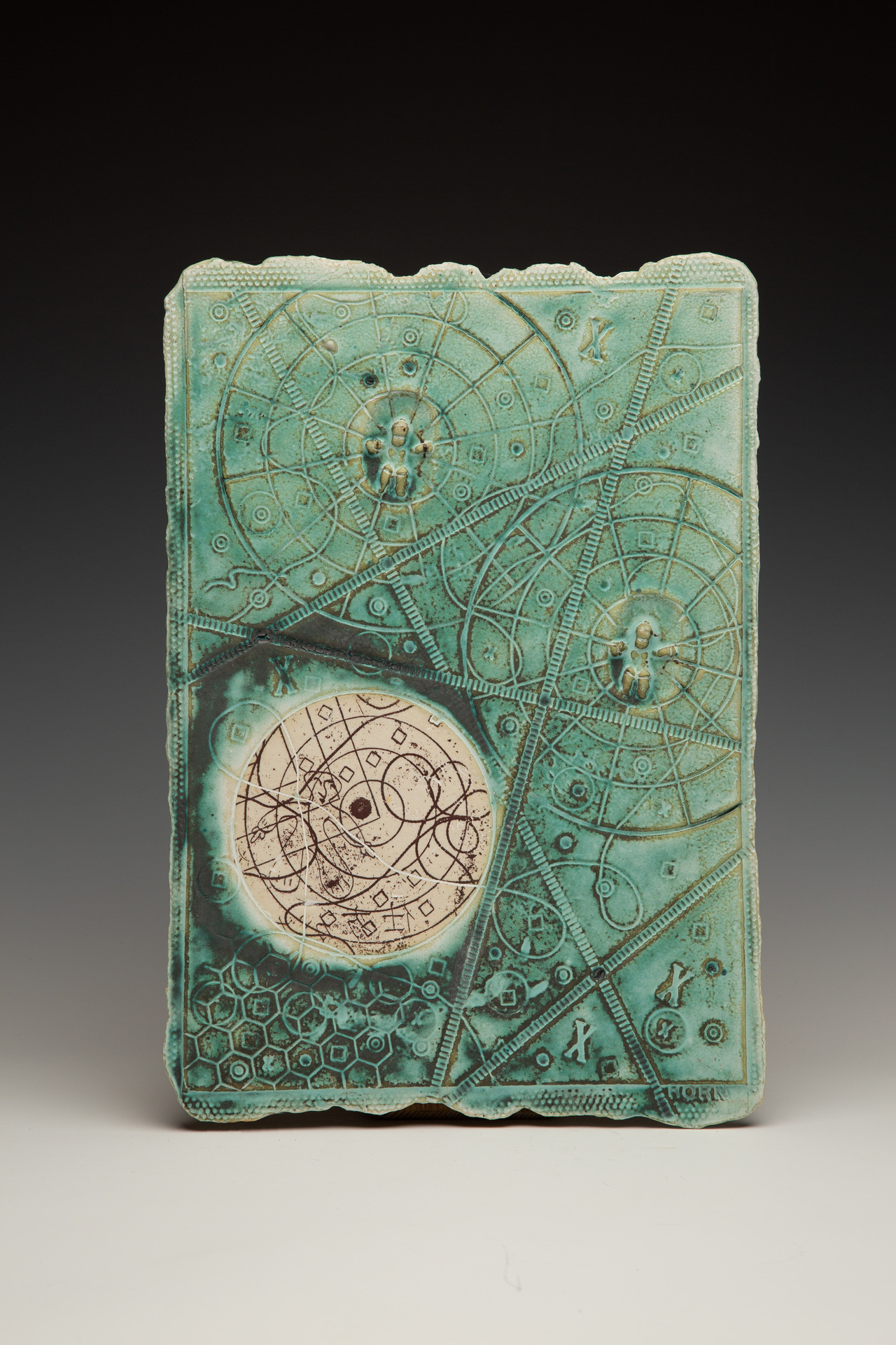 A rectangular ceramic piece in a turquoise color featuring intricate circular patterns and geometric designs. It includes a central cream-colored circle filled with abstract symbols and several raised human figures within the circular motifs.