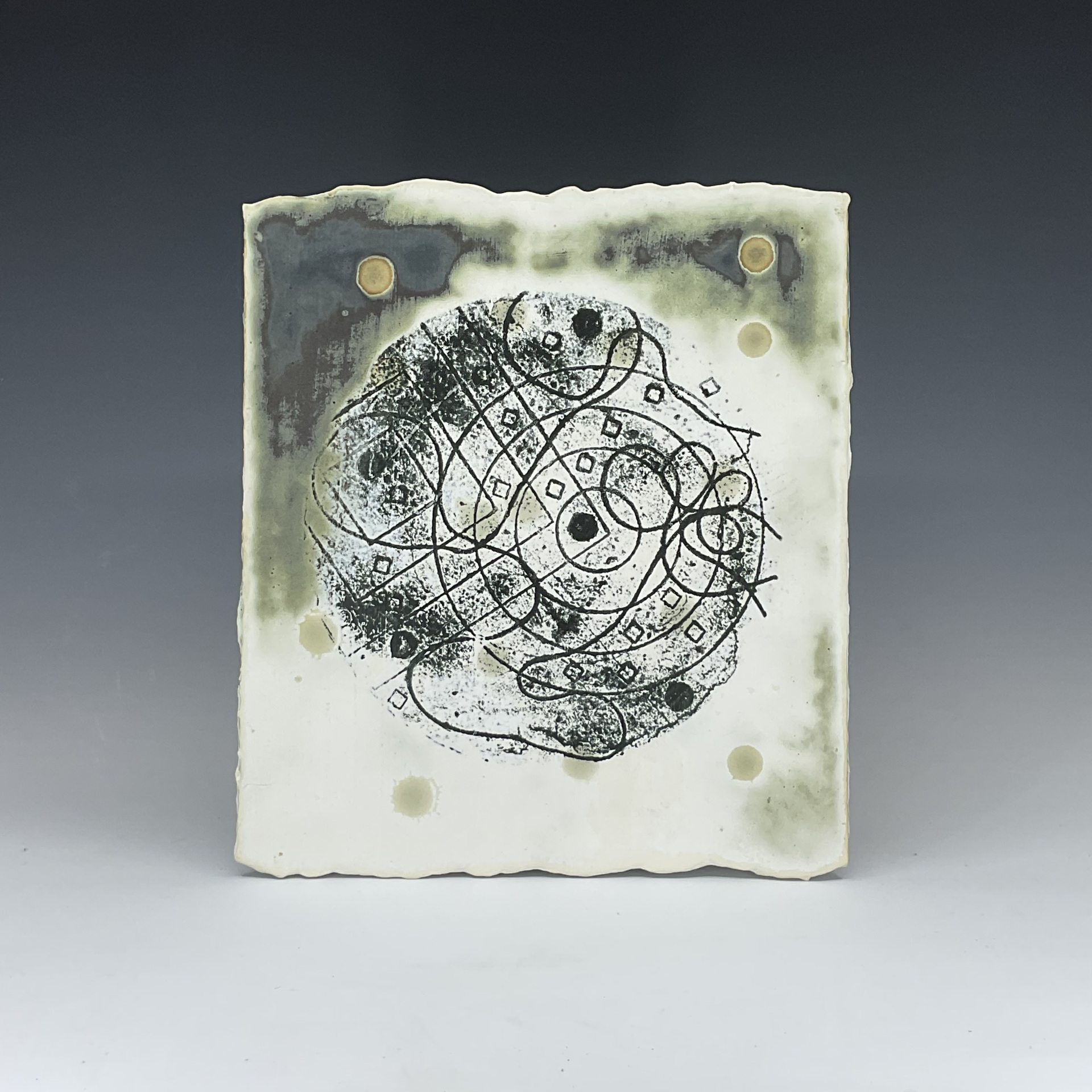 A square ceramic tile with an abstract black circular pattern in the center, surrounded by soft green and cream tones. The tile has an uneven edge, adding to its organic feel, with small circular impressions scattered throughout.