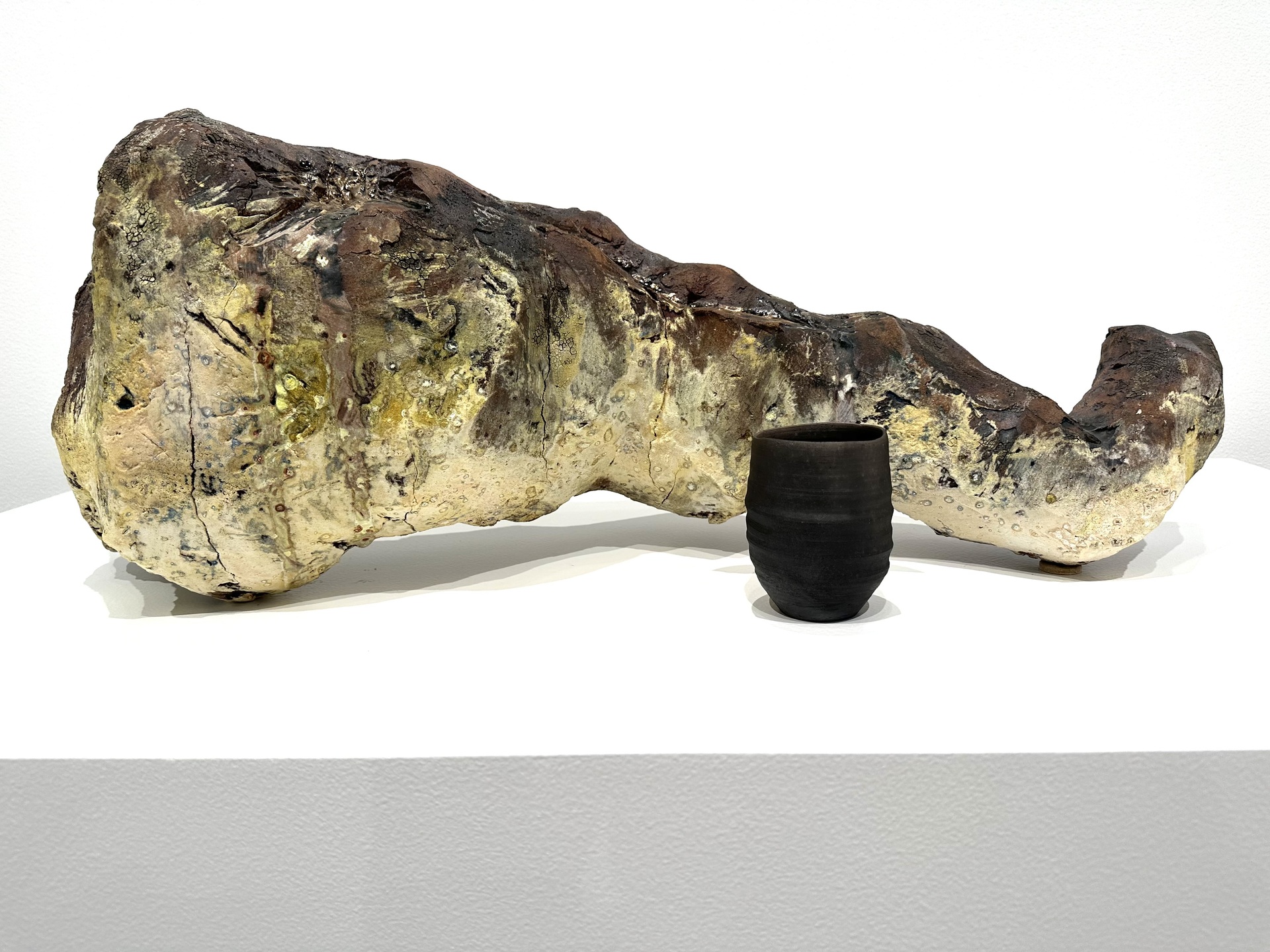 A large, abstract ceramic sculpture with a rugged, earth-toned texture resembling natural rock, displayed next to a small, dark cup-shaped vessel on a white platform.