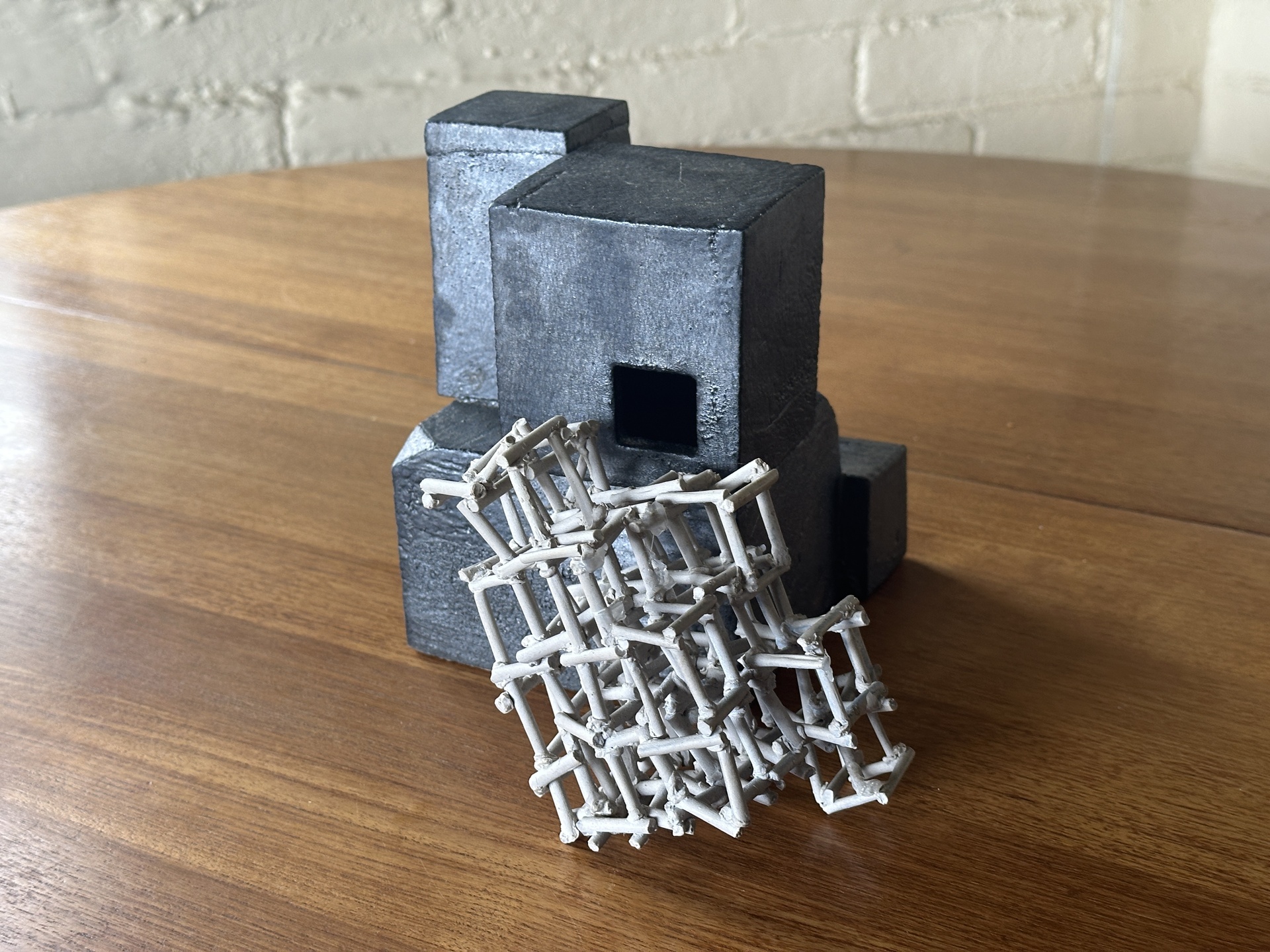 A sculpture featuring geometric black blocks with hollow sections, paired with a tangled, lattice-like white structure resting against them, placed on a wooden surface.