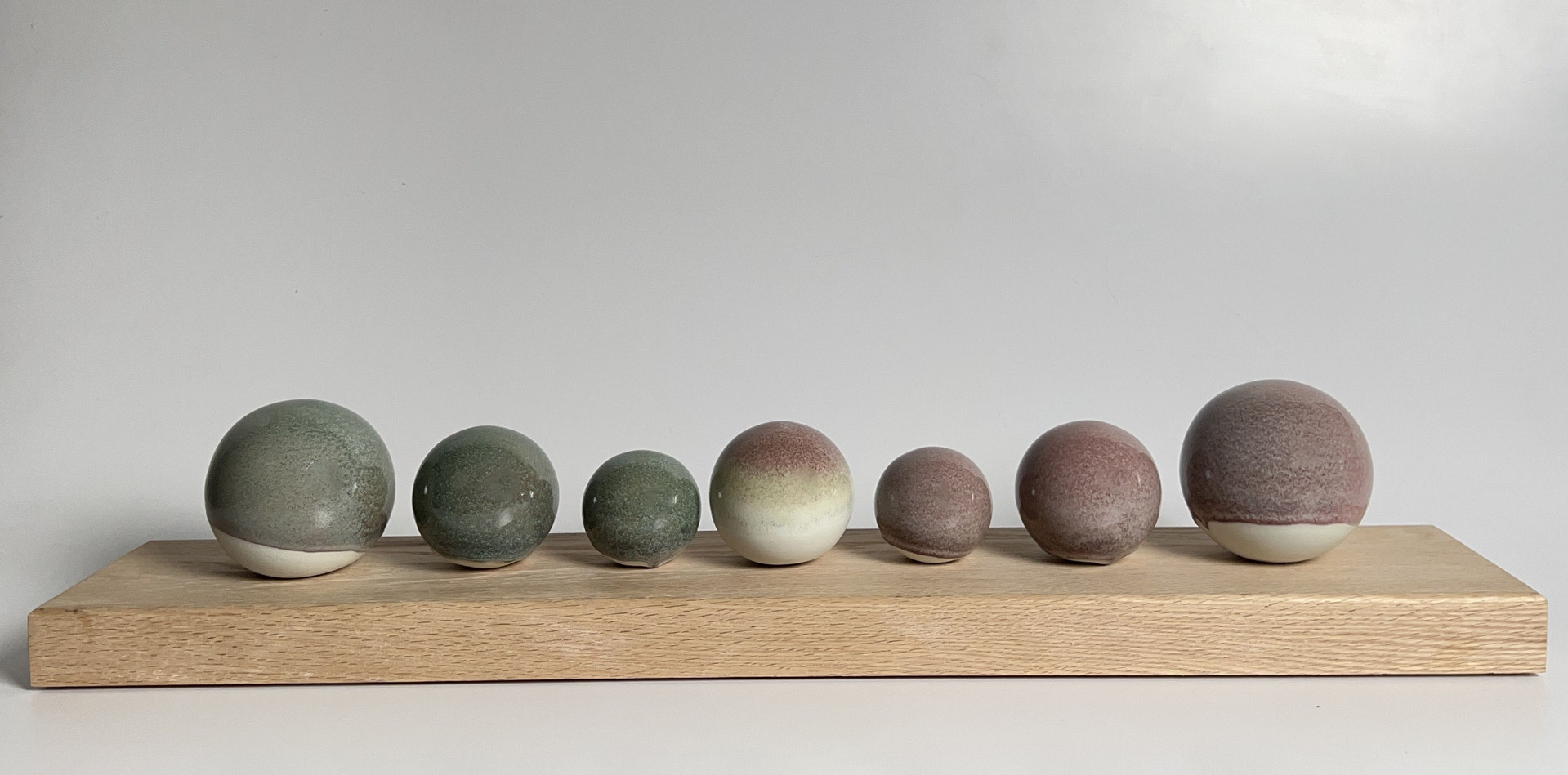 A series of spherical ceramic objects arranged in a row on a wooden base, gradually increasing in size from left to right, with surfaces in muted shades of green and reddish-brown.