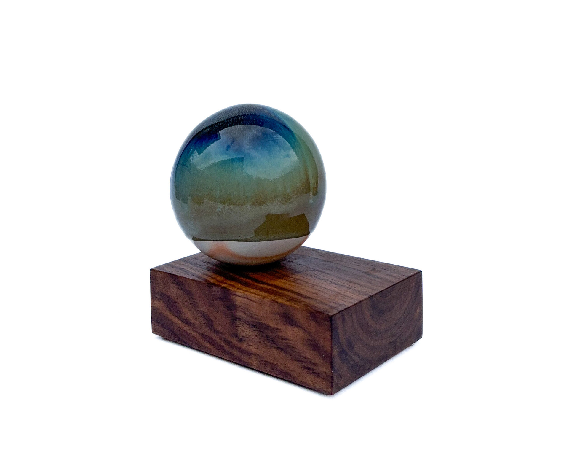 A glossy spherical ceramic object with a gradient of earthy and blue tones, placed on a smooth wooden block, creating a contrast between the polished sphere and the natural grain of the wood.