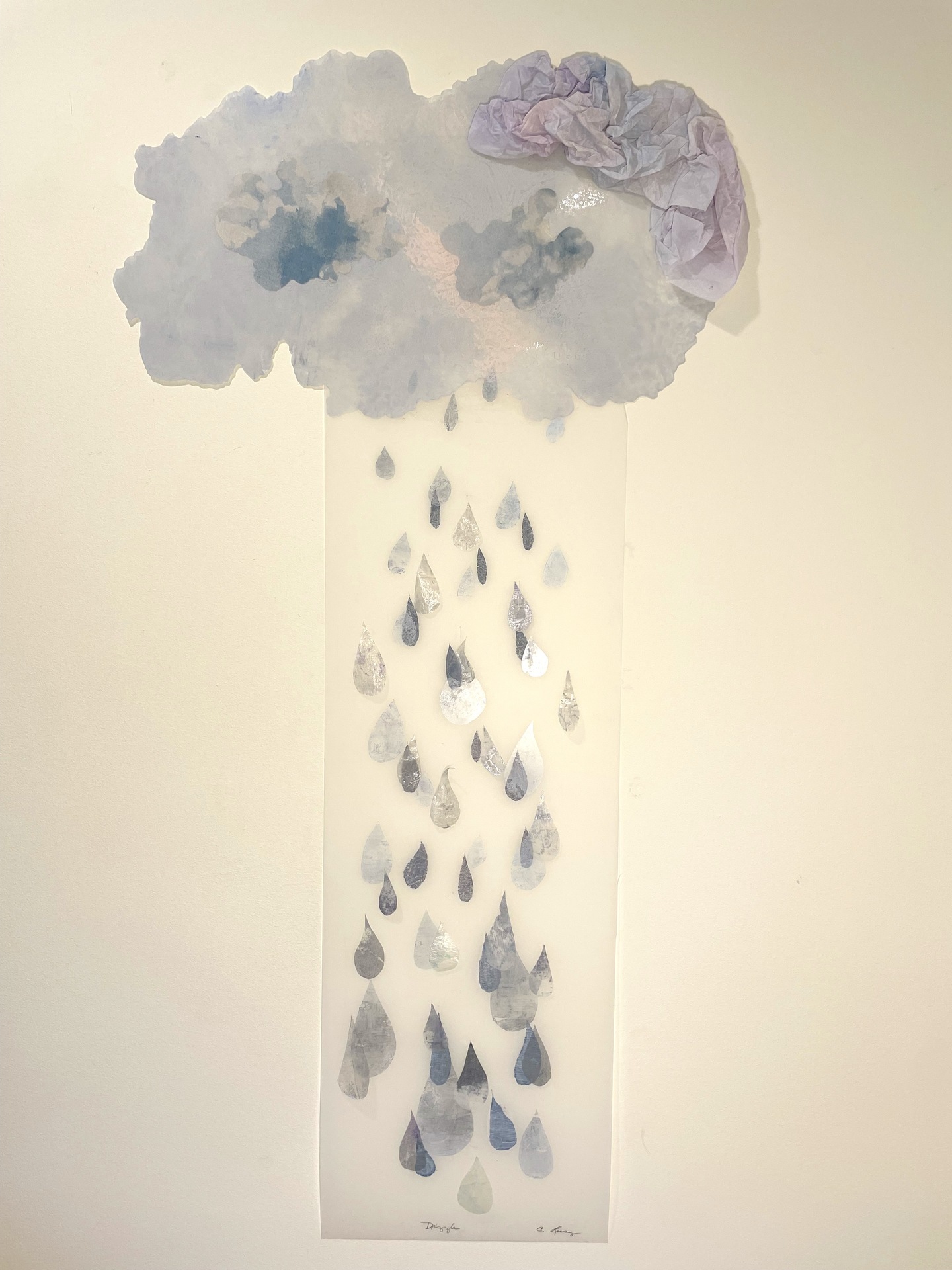 A mixed-media artwork depicting a cloud with soft, textured edges and crumpled material in shades of gray and purple, releasing raindrop-shaped cutouts in various shades of blue, cascading downward.