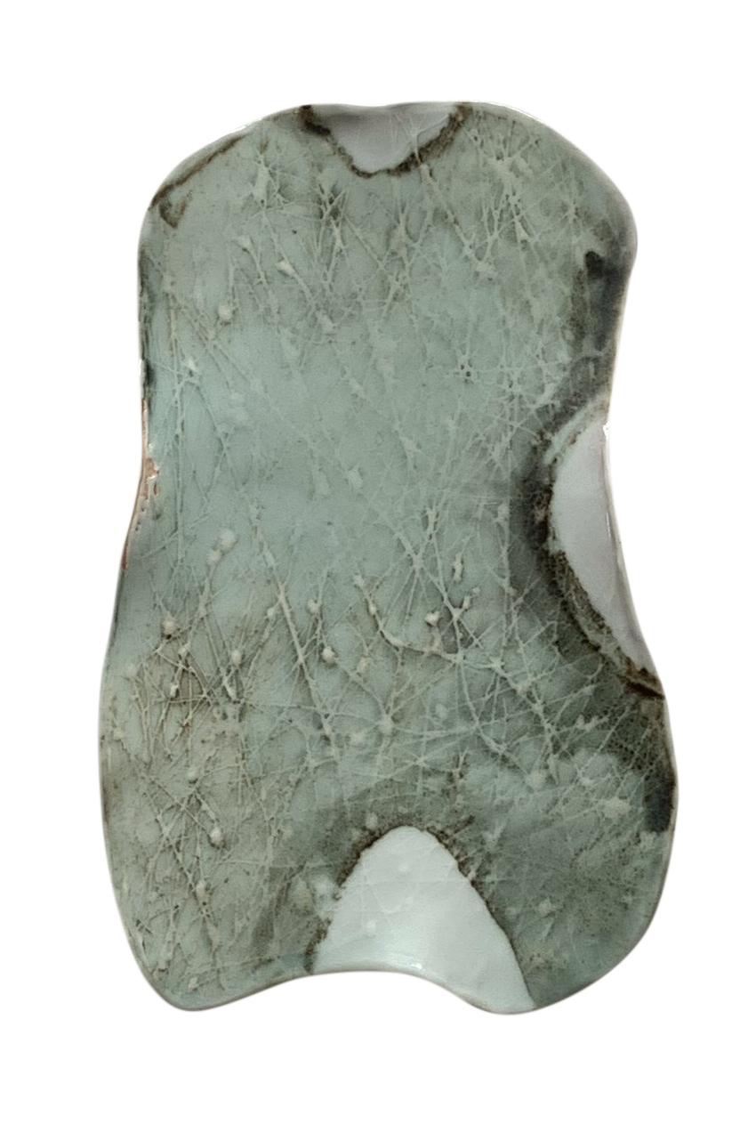 An abstract ceramic piece with an irregular form, featuring a soft green surface marked by intricate crackling and textural details, accented with white patches that create an organic, weathered appearance.