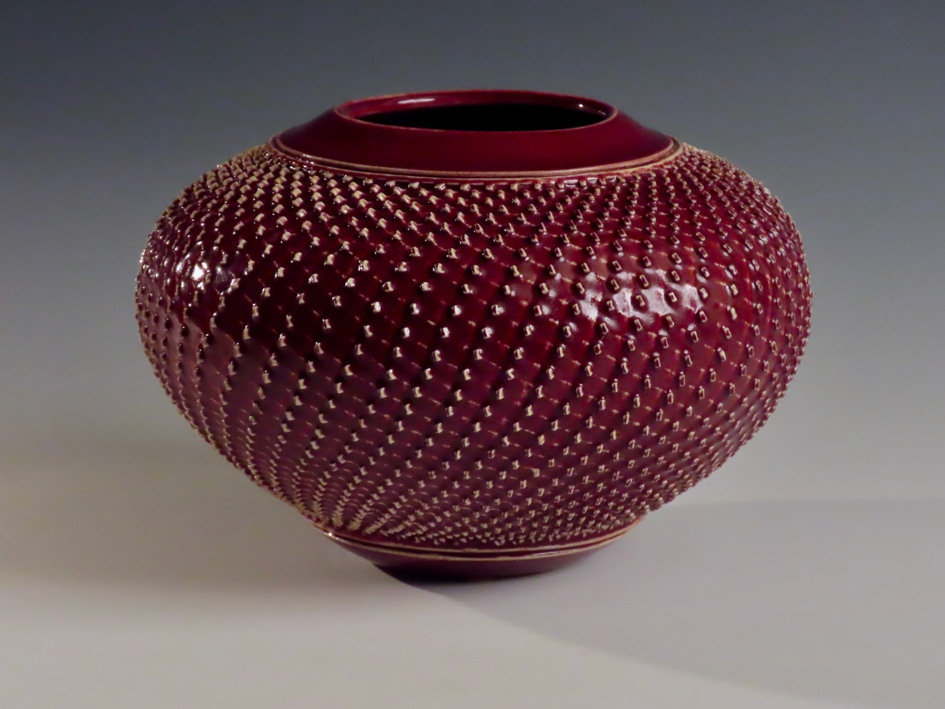 A deep red ceramic vase with a round, bulbous shape, featuring a surface covered in evenly spaced, textured bumps that create a patterned, tactile effect.