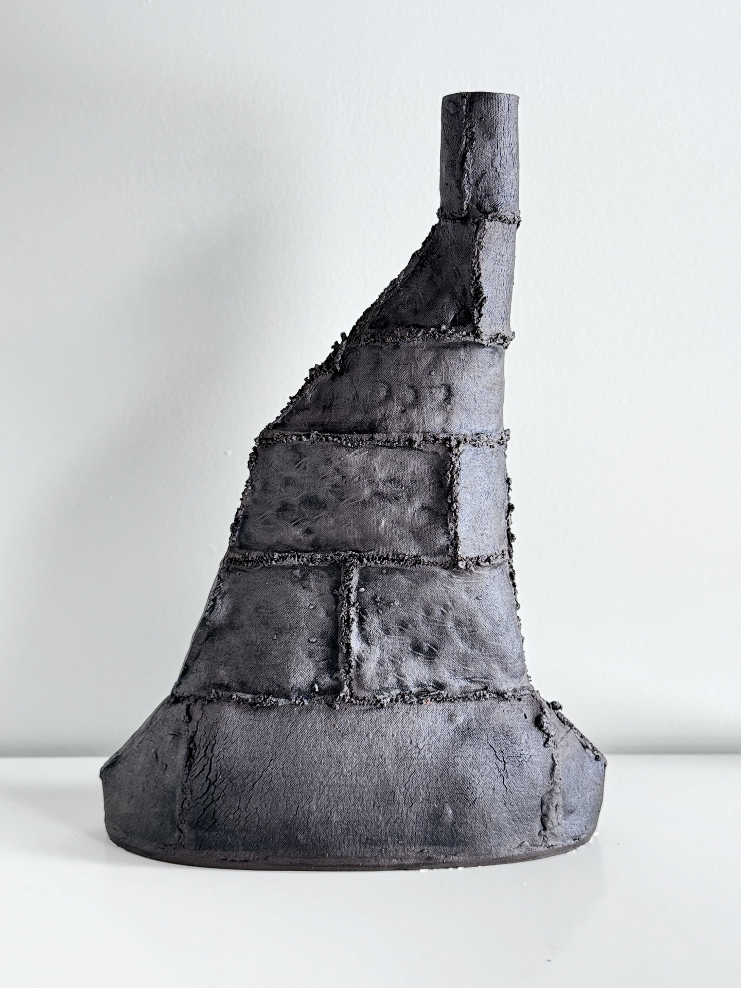 A dark, textured ceramic sculpture with an asymmetrical, conical form, featuring segmented, brick-like sections and a narrow, cylindrical top, giving it a rugged, industrial appearance.