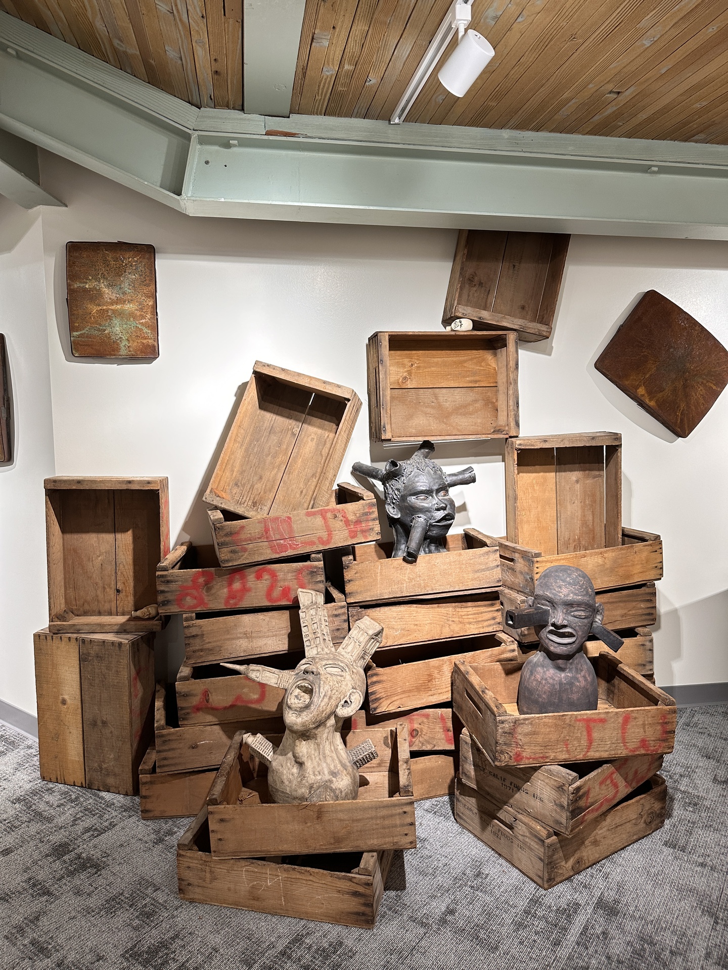 A display of wooden crates stacked in an irregular, leaning manner, with several crates arranged at different angles. Inside some of the crates are sculptures resembling ancient or tribal head figures. The sculptures have exaggerated facial expressions: one has an open mouth, possibly mid-yell, and another has large cylindrical shapes protruding from its face, creating an intense and dramatic appearance. The crates are weathered, with visible signs of wear, and some have red markings or stenciled numbers on the outside.