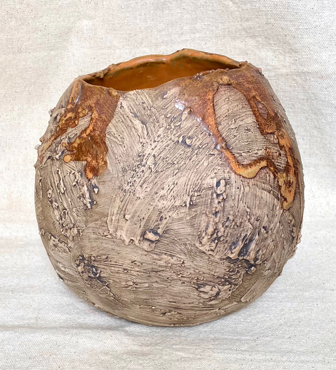 A handcrafted ceramic vessel with a rounded, bulbous shape and a rough, textured surface. The exterior of the vessel is predominantly earthy brown, with large, sweeping strokes of clay creating a natural, rustic look. Some areas near the top feature a reddish-brown glaze that appears to have dripped down organically, forming abstract patterns that contrast with the matte, unglazed surface of the body.