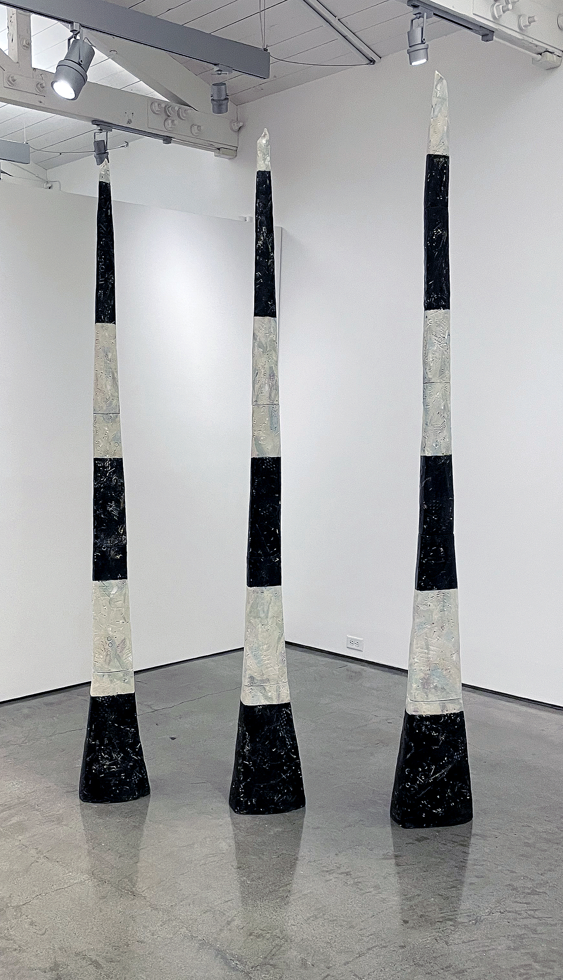 Three tall, cone-shaped sculptures with alternating black and white sections standing in a gallery space.