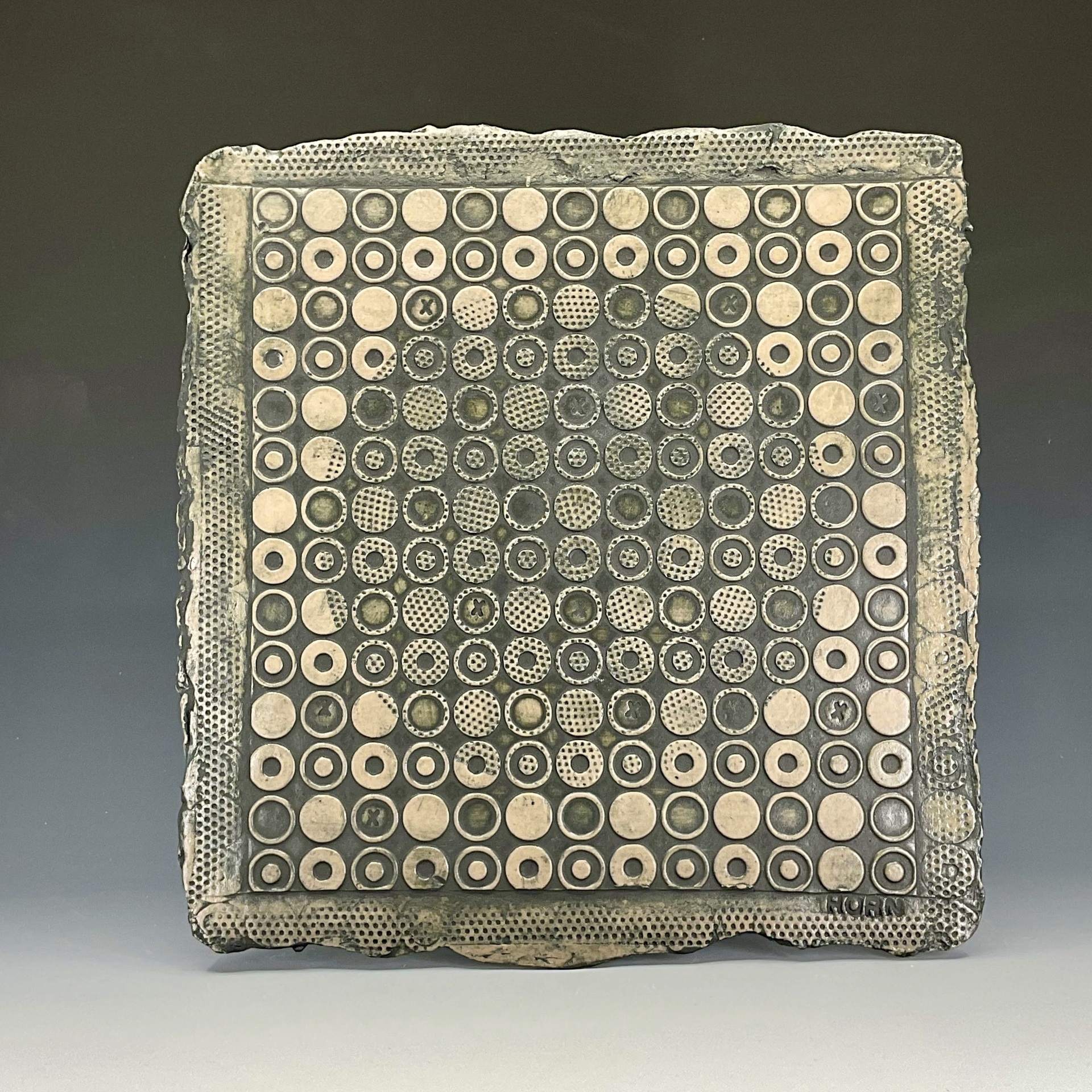 A square ceramic tile featuring a grid of various circular patterns, with raised textures and repeating designs in earthy tones, framed by a slightly rough, uneven edge.