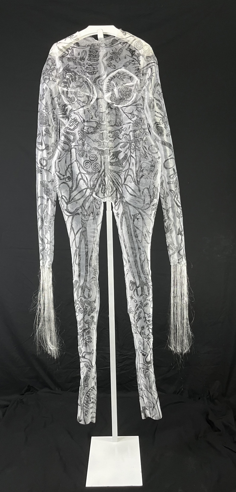 A sheer, full-body garment decorated with intricate black illustrations of various organic forms, displayed on a white stand against a black background, with long, delicate strands extending from the sleeves.