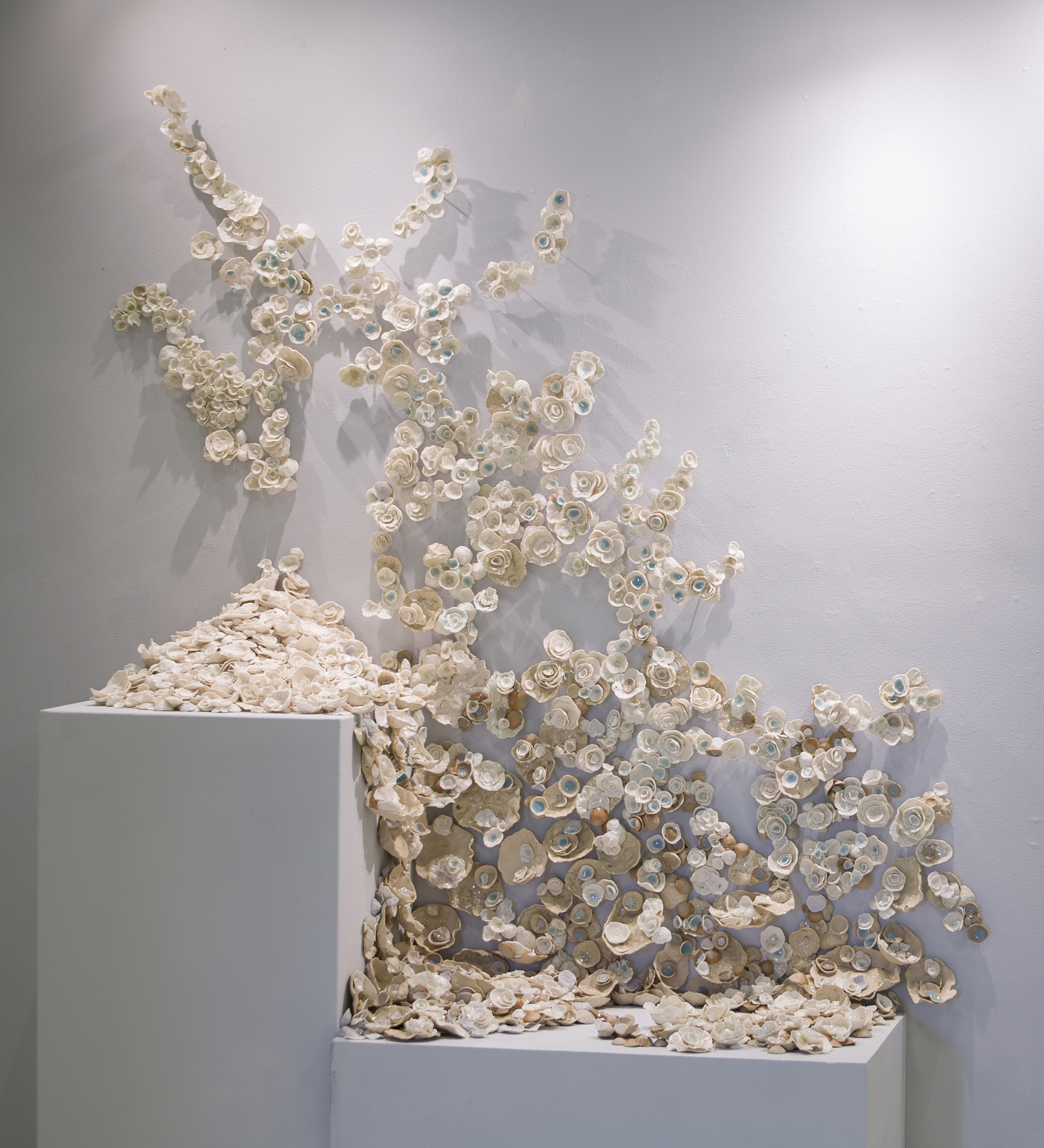 A wall-mounted installation featuring an arrangement of delicate, cream-colored ceramic rosettes, some with light blue centers, cascading from two white pedestals and spreading organically across the surface, evoking the appearance of blooming flowers or coral.