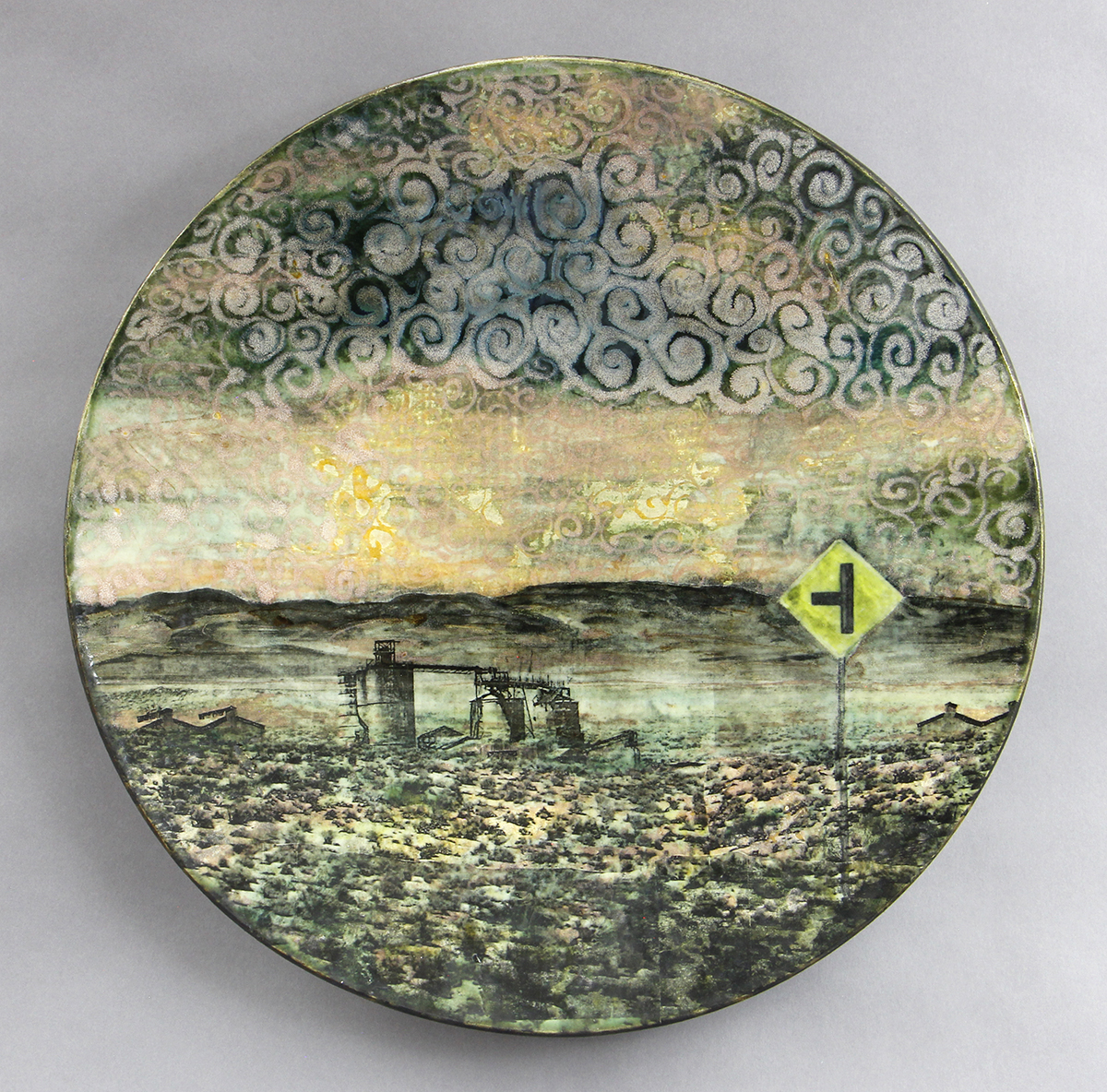 A round ceramic piece depicting a surreal landscape with swirling clouds, a desert scene with abandoned structures, and a road sign suggesting an intersection ahead.