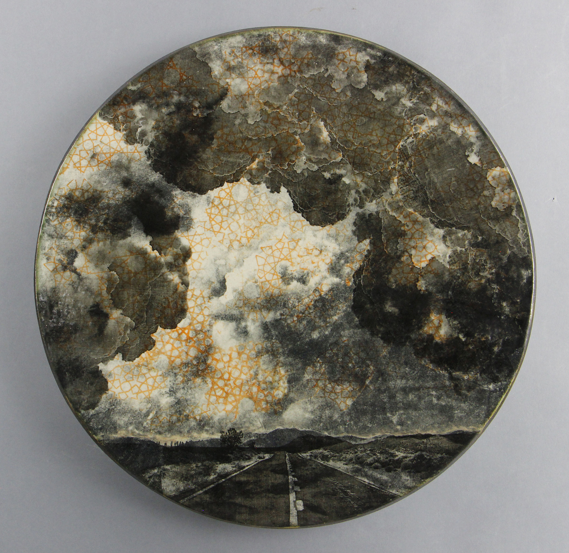 A round ceramic piece featuring a dark, dramatic sky with clouds over a desert road, highlighted by a subtle geometric pattern interspersed within the clouds.