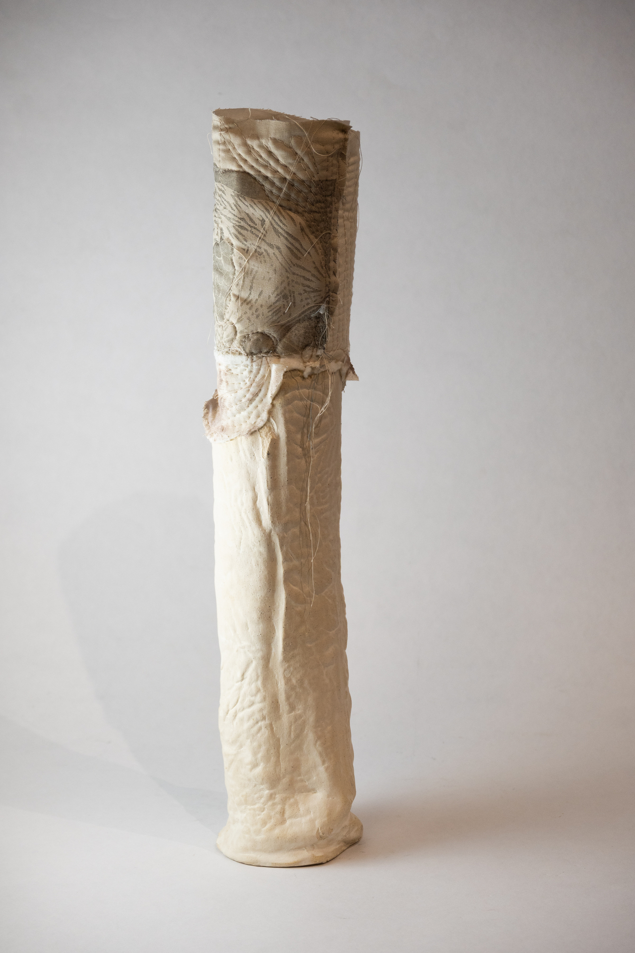 A tall, cylindrical sculpture with a quilted texture and stitched fabric elements in light and neutral tones.