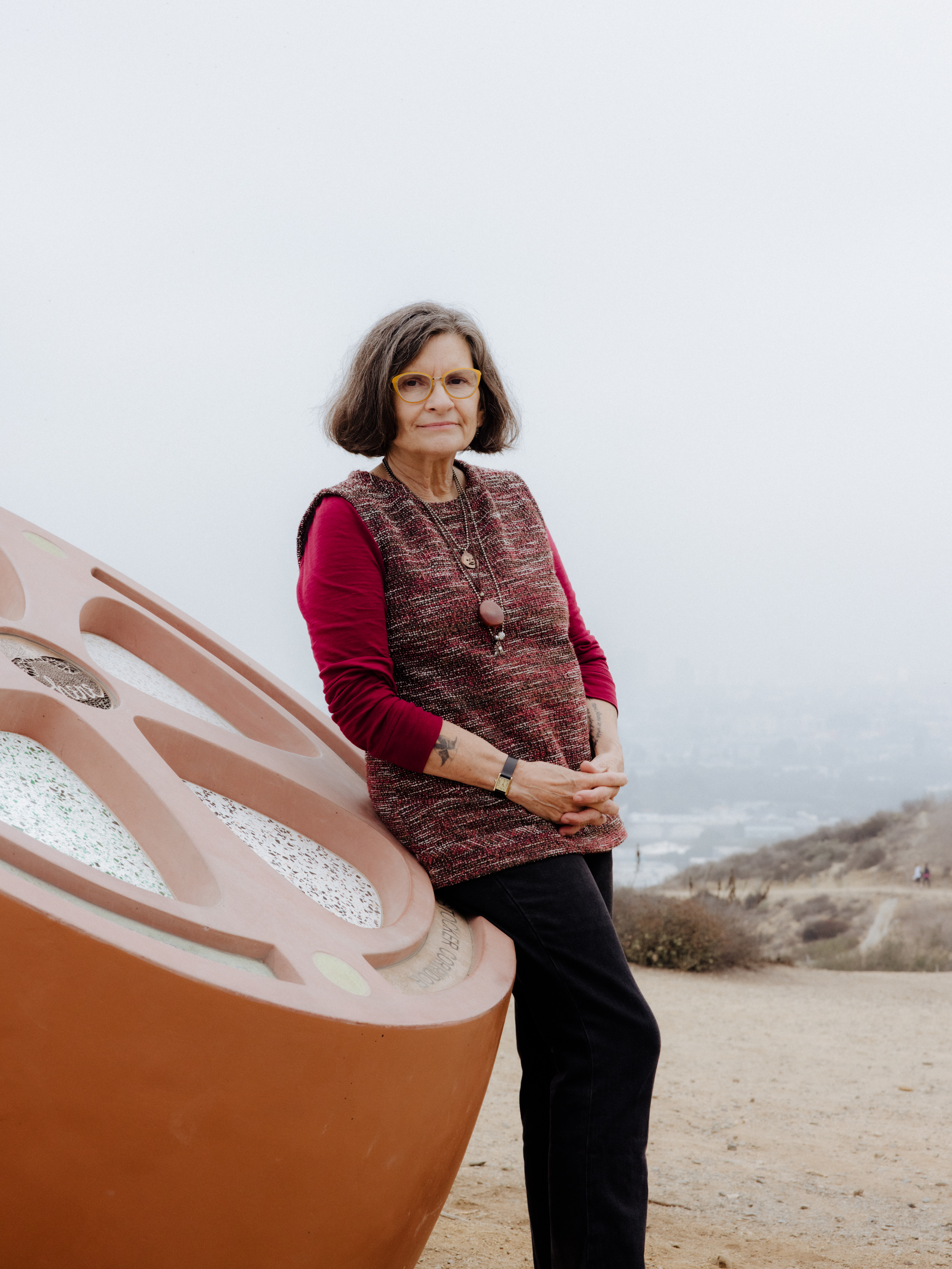 Kim Abeles, she is on a hilltop leaning on an orange sculpture while wearing yellow glasses, a red patterned overshirt with a red longsleeve underneath. Photography by Joyce Kim