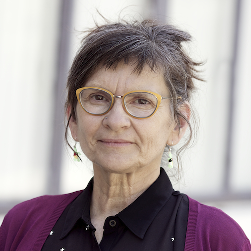 Kim Abeles, she is standing in front a white blurred background while wearing yellow glasses, a purple cardigan and a black buttoned shirt. Photography by Calista Lyon